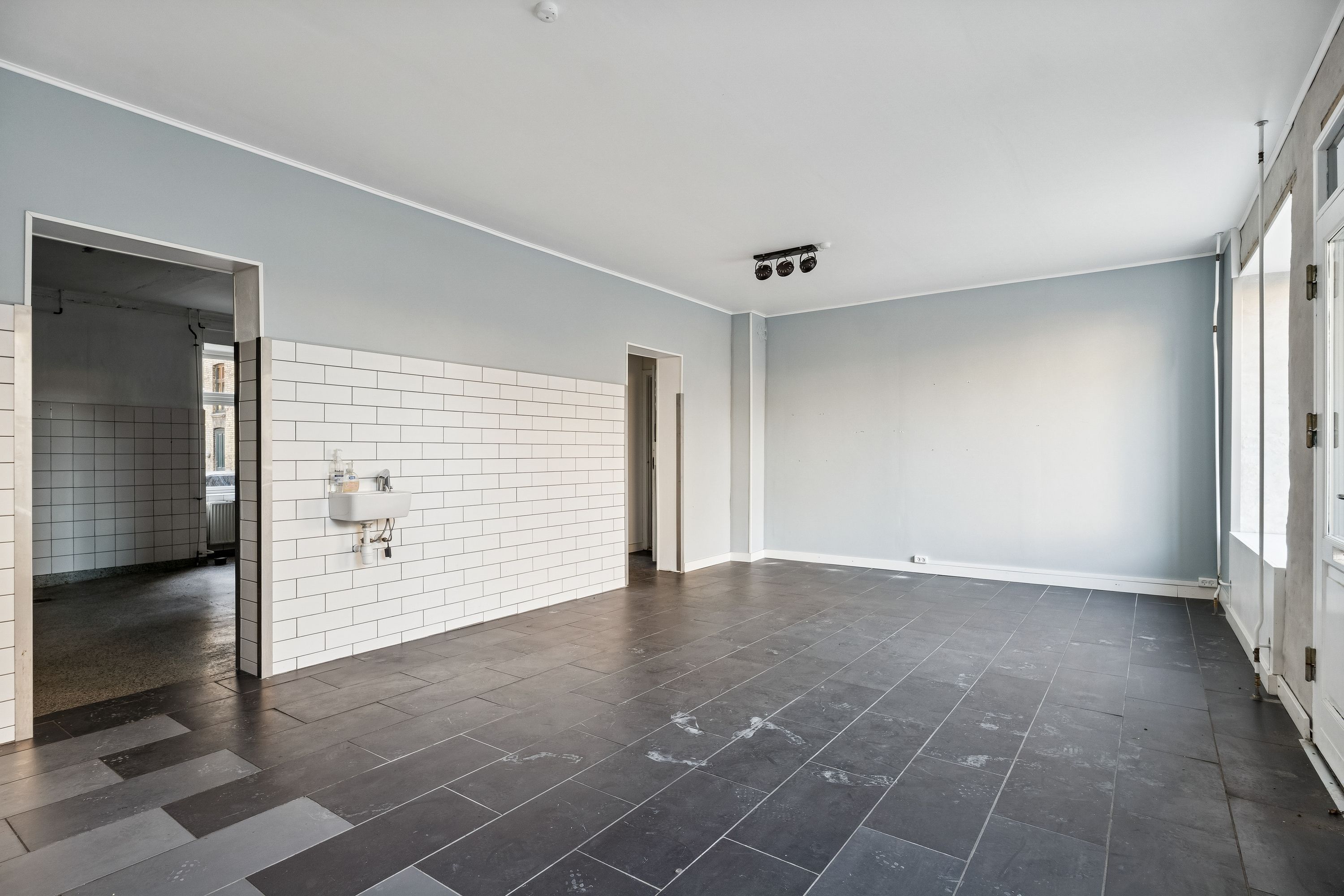 En tom stue med lysegrå vægge og sortgule klinker på gulvet og en hvid vask med vandhaner mod en dør.