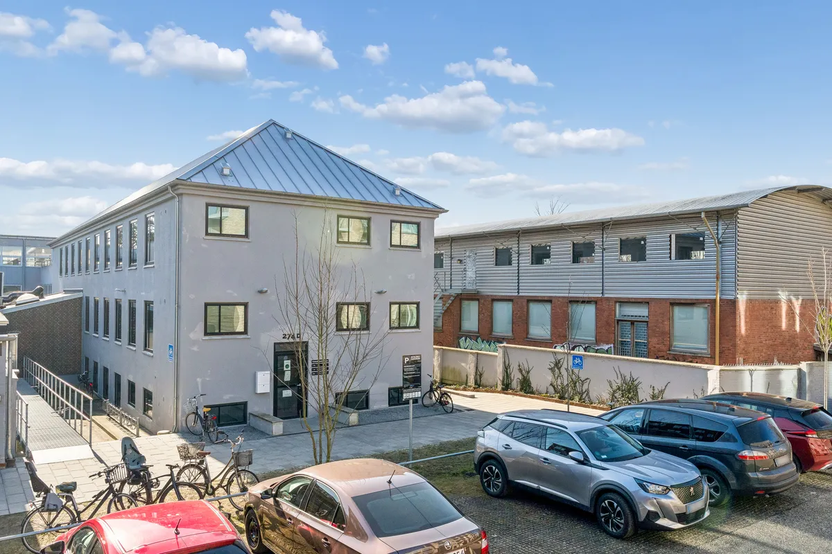 Kontorlokaller på Frederikssundsvej 274D i Brønshøj med parkeringsplads og udsigt til Frederikssundsvej og Husumvej. Mange biler parkeret og to cykler stående på cykelstel.