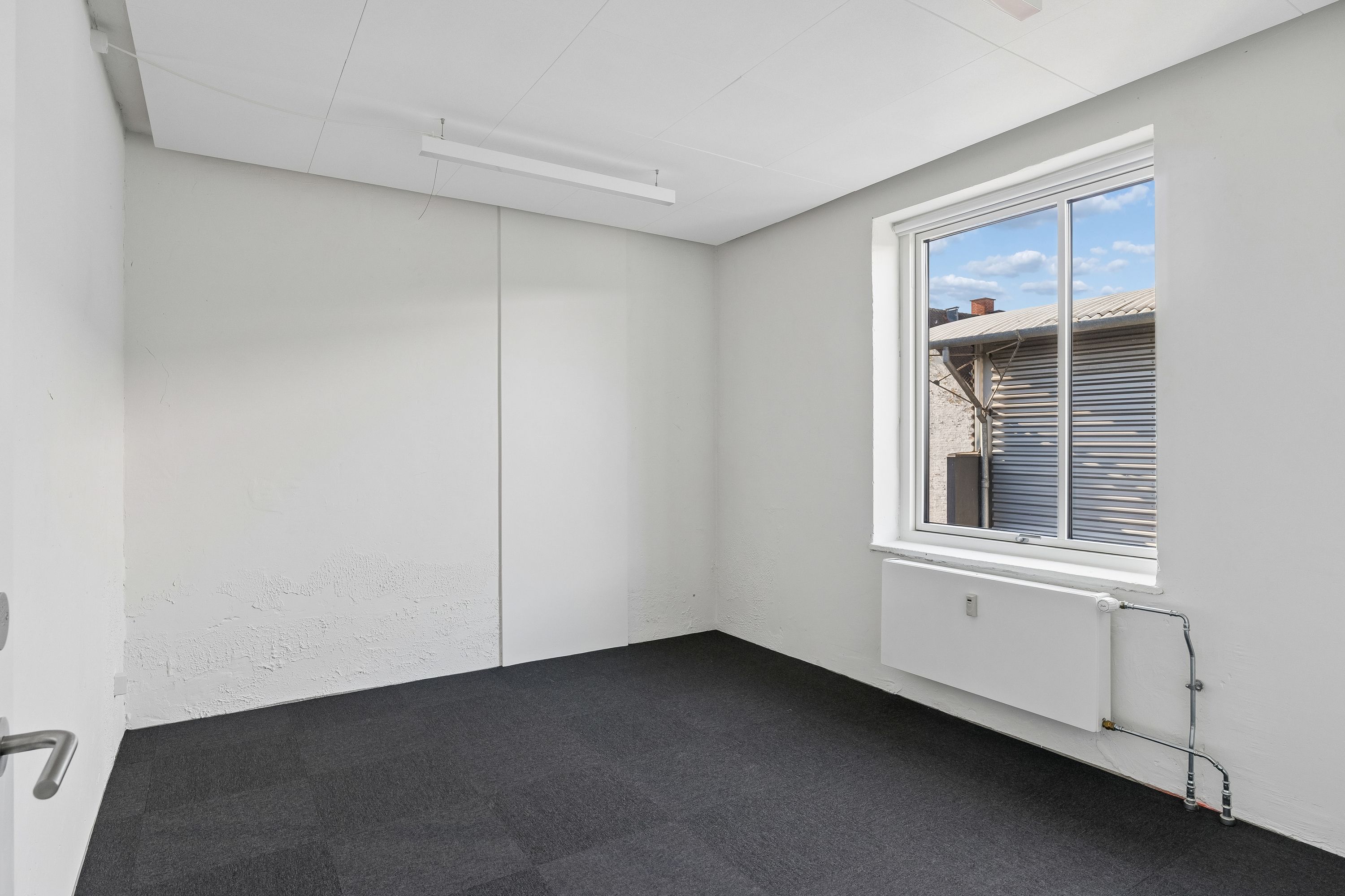 Kontor til leje i Brønshøj med lyse kontorlokaler, moderne radiatorer og vinduer med udsigt til bygninger og en røgskorsten. Lokalerne har hvide vægge og loft og er fuldstændig nye.