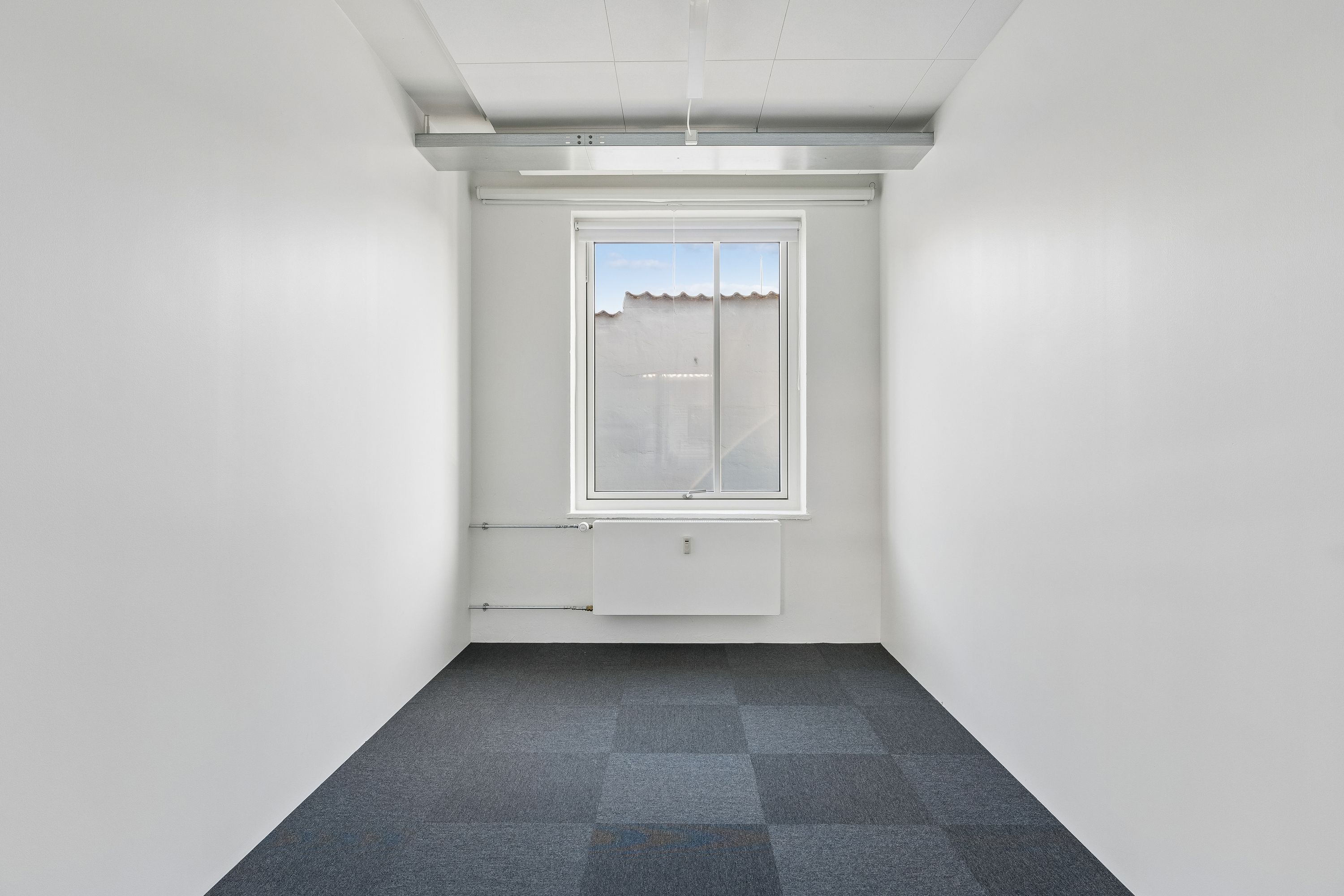 Rummelig og lyse kontorlokaler med hvidt vægpanel og møbler. Store vinduer med gardiner og radiator under. Moderne hvide loftsbjælker og gulvbelægning.