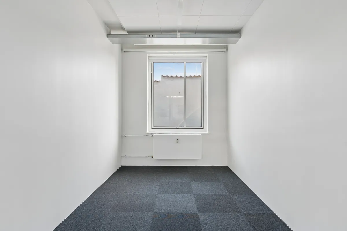 Rummelig og lyse kontorlokaler med hvidt vægpanel og møbler. Store vinduer med gardiner og radiator under. Moderne hvide loftsbjælker og gulvbelægning.