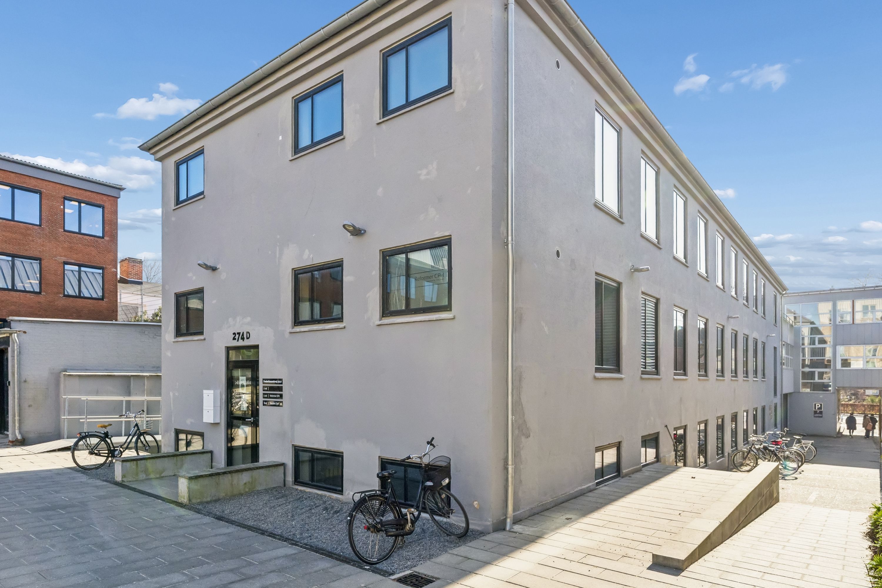Kontor til leje på Frederikssundsvej 274D i Brønshøj. Kontorbyggeriet med flere vinduer og to cykler parkeret foran. Lys himmel med få skyer.