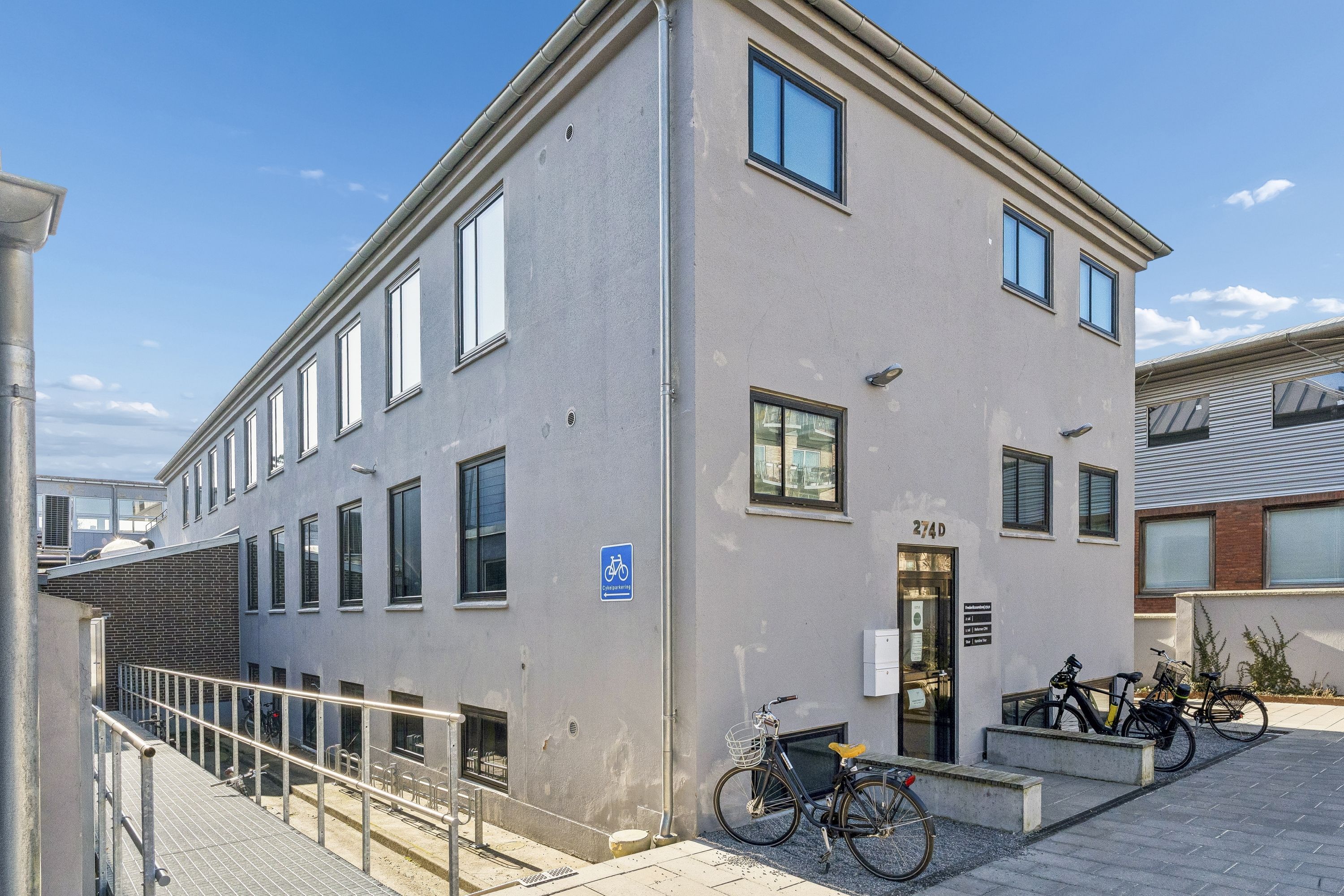 Kontorlokaller med glasruder og moderne indretning, liggende på Frederikssundsvej. Cykler parkeret foran bygningen. Annonce for kontorlokaler og kontorer på 2. sal.