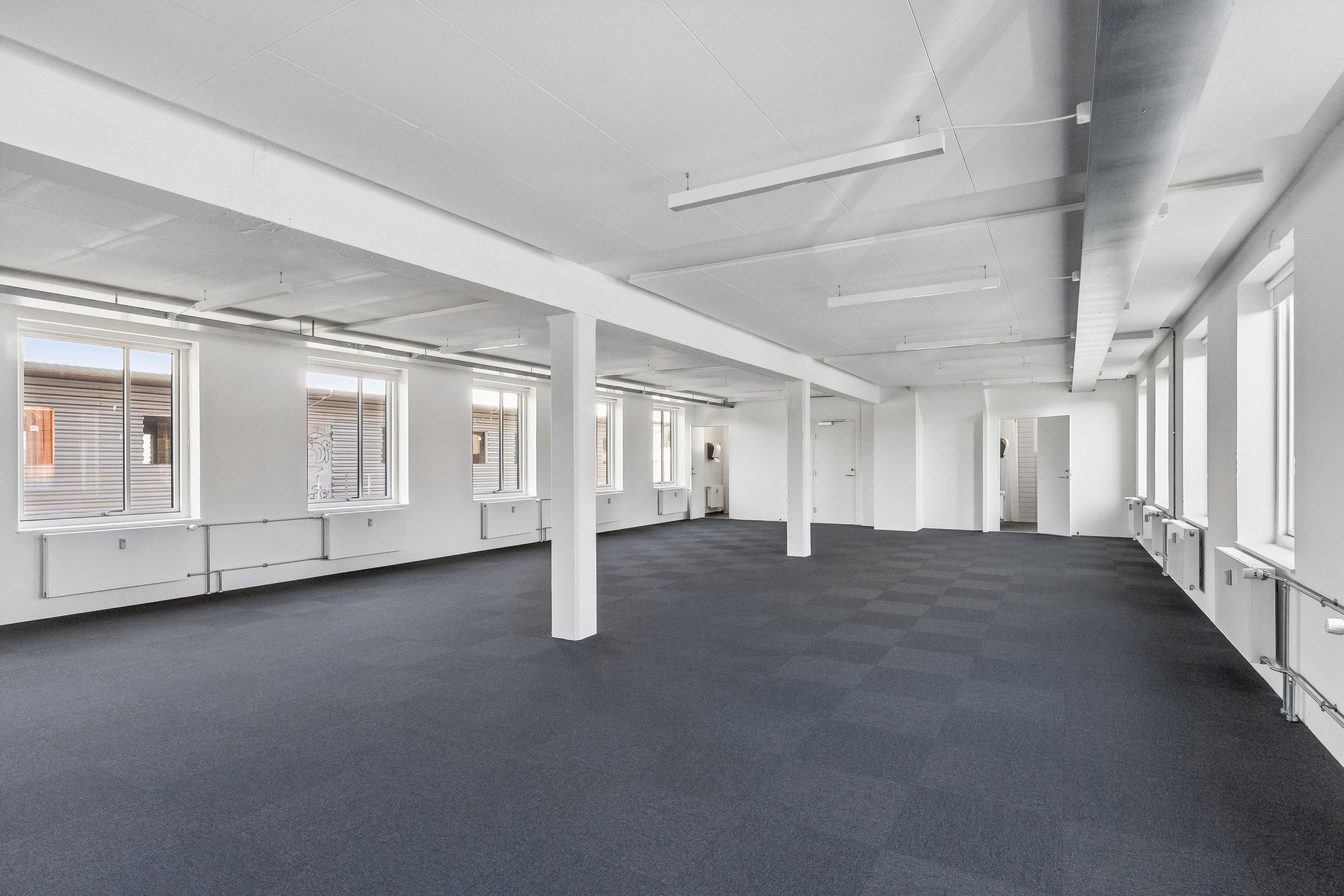 Foto af et tomt og lyse kontorruum med hvide vægge, møbler, vinduer, gulv og loftslampe. De store vinduer er åbne, og der er radiatorer og håndtag på væggene.