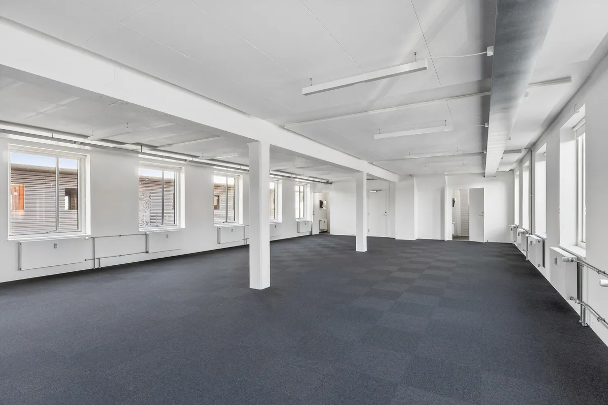 Foto af et tomt og lyse kontorruum med hvide vægge, møbler, vinduer, gulv og loftslampe. De store vinduer er åbne, og der er radiatorer og håndtag på væggene.