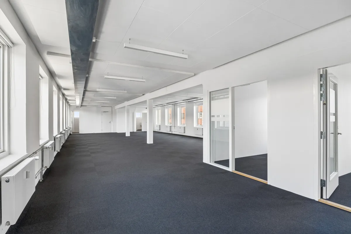 Foto af et tomt kontorrum med hvide vægge, sort gulv, store vinduer og flere døre. Der er lysekroner og radiatorer på væggene