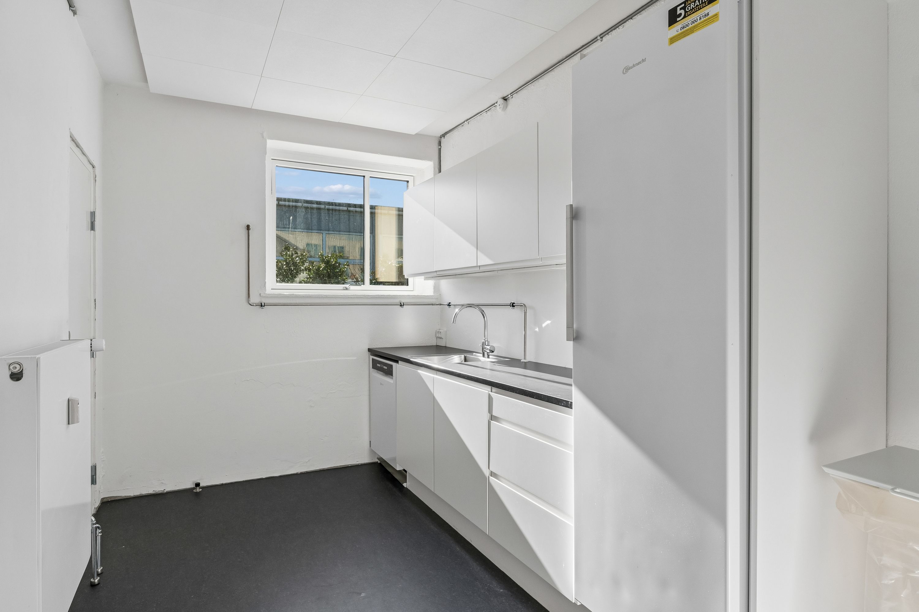 Kontor med hvidt loft, mørk gulvbelægning og hvide vægge. Et vindue med udsigt til en bygning og en plante samt en håndvask med vandhane.