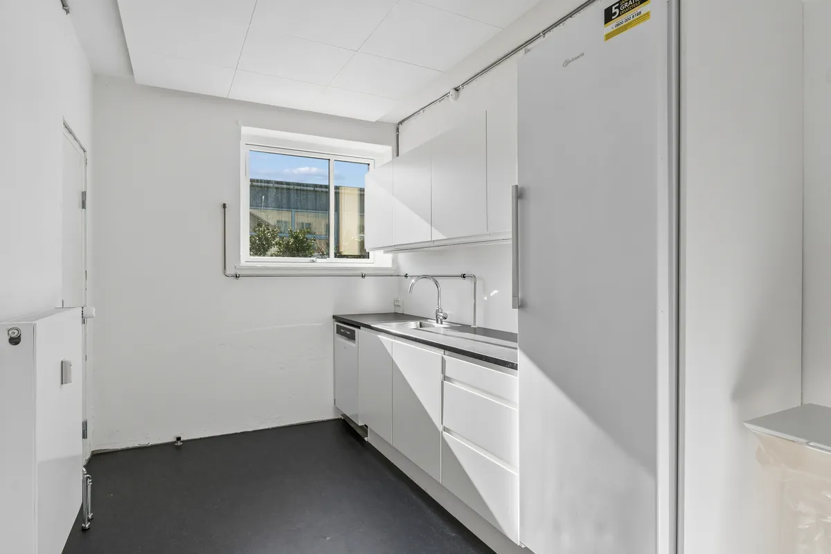 Kontor med hvidt loft, mørk gulvbelægning og hvide vægge. Et vindue med udsigt til en bygning og en plante samt en håndvask med vandhane.