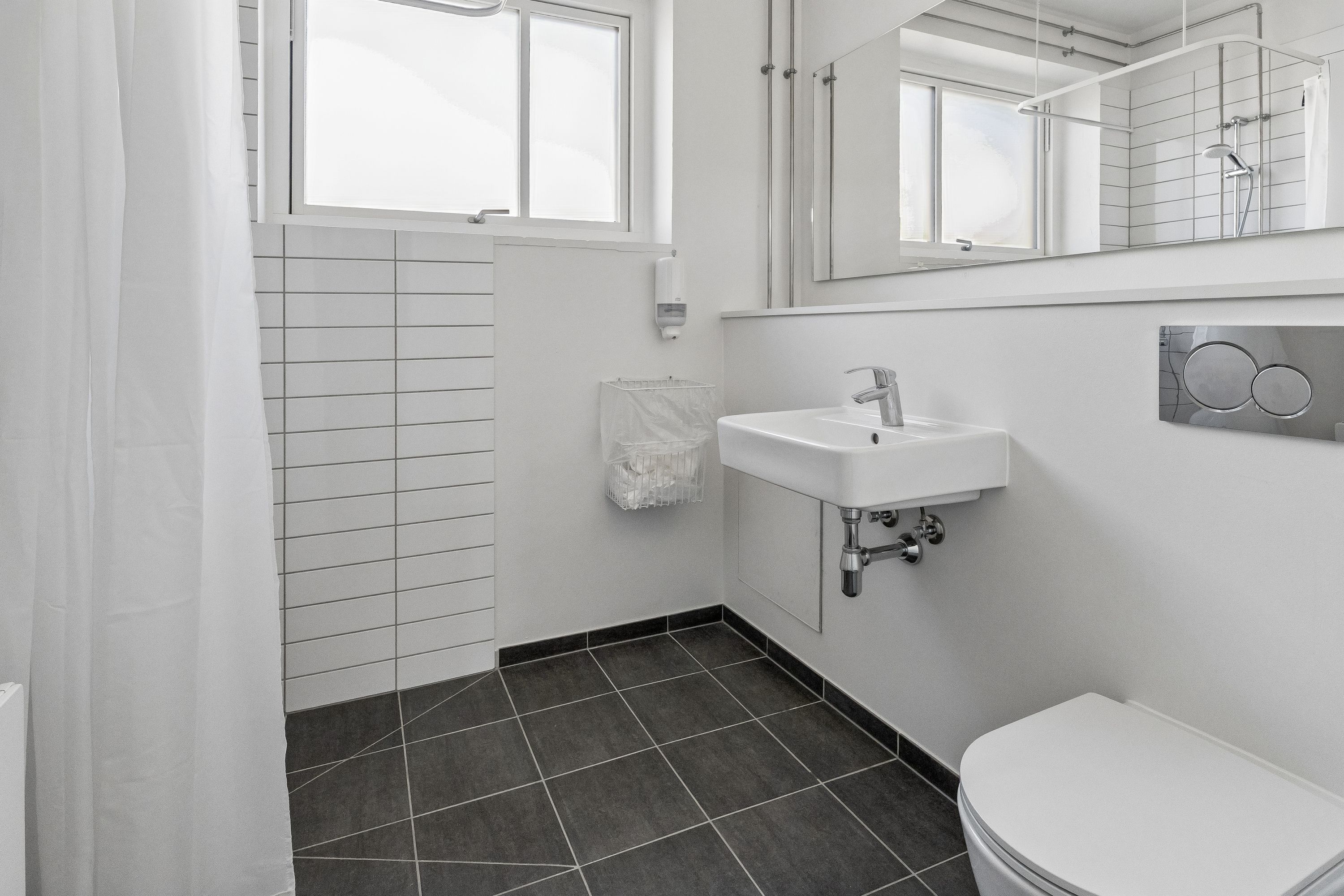 Rent kontor med hvidt interiør, toalett, håndvask, vinduesruder og håndklædeholder. Toilettet er hvidt og ligger ved siden af håndvasken og spejlet