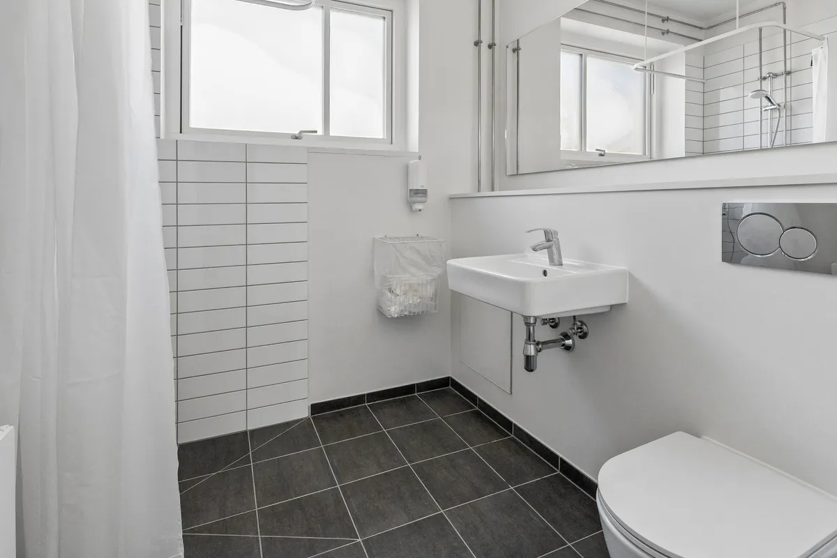 Rent kontor med hvidt interiør, toalett, håndvask, vinduesruder og håndklædeholder. Toilettet er hvidt og ligger ved siden af håndvasken og spejlet