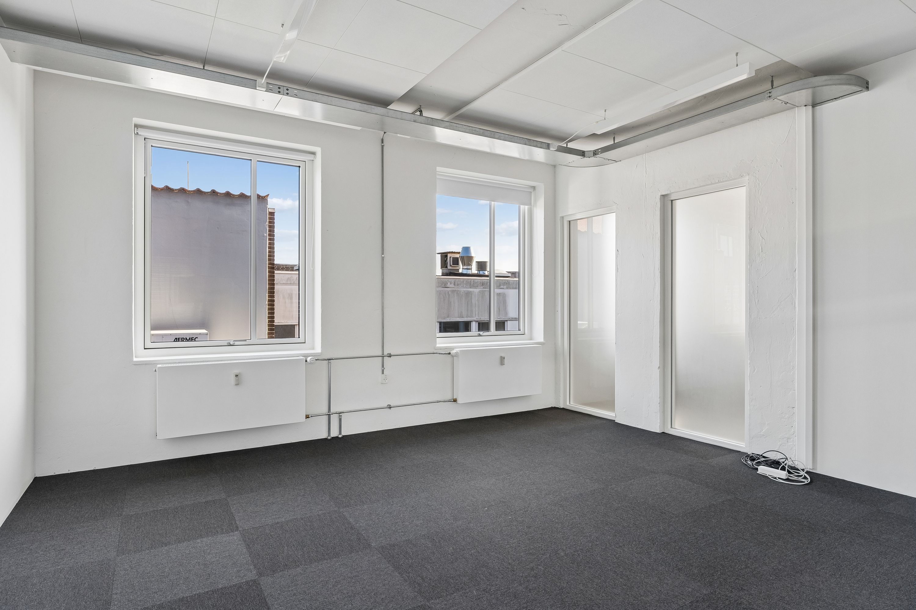 Lækre kontorlokaler med lyseruder og hvide vægge, gulvtæppe og moderne radiatorer, krydsfinerloft og teknisk udstyr samt 2 vinduer med udsigt til gaderne og bygninger
