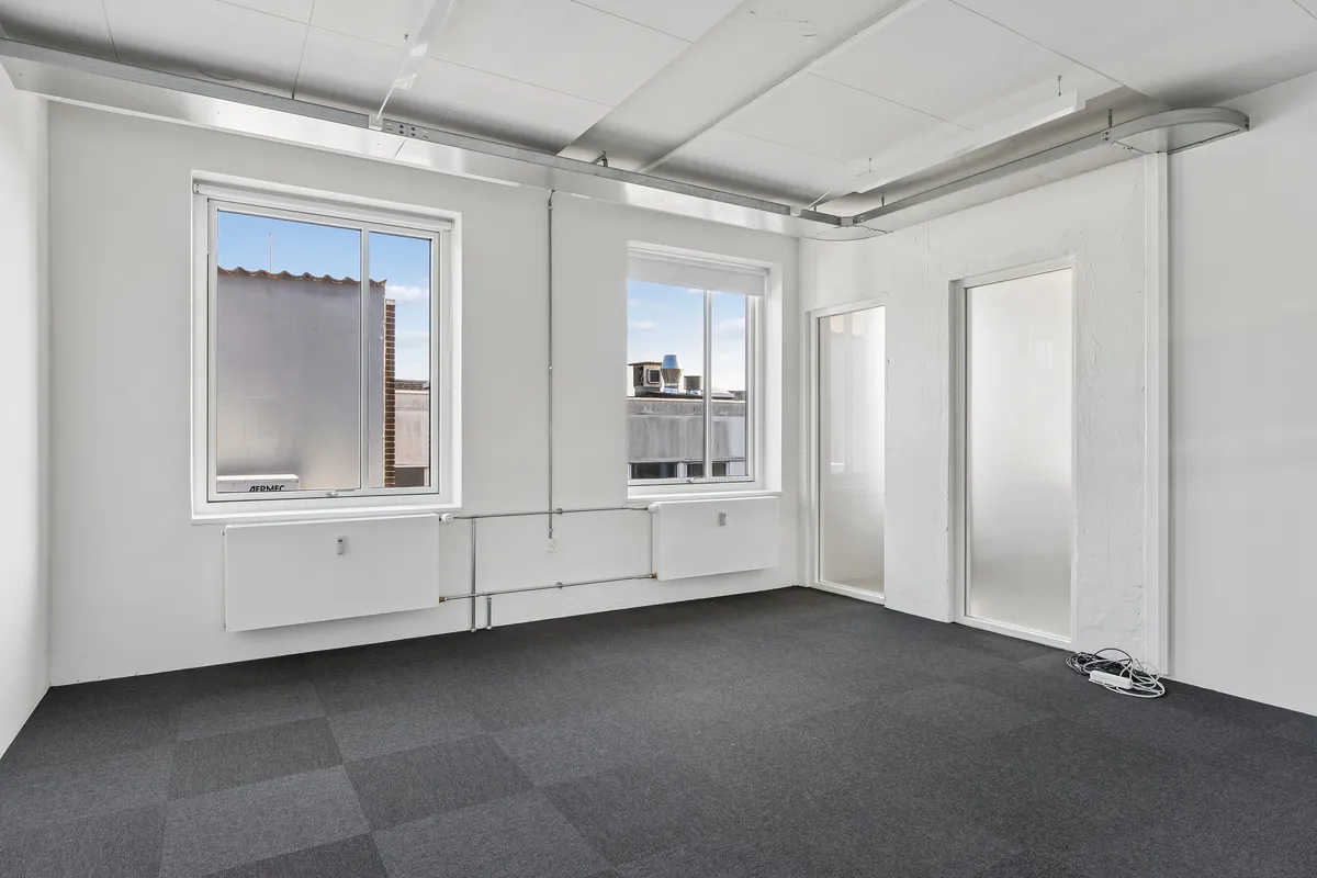 Lækre kontorlokaler med lyseruder og hvide vægge, gulvtæppe og moderne radiatorer, krydsfinerloft og teknisk udstyr samt 2 vinduer med udsigt til gaderne og bygninger