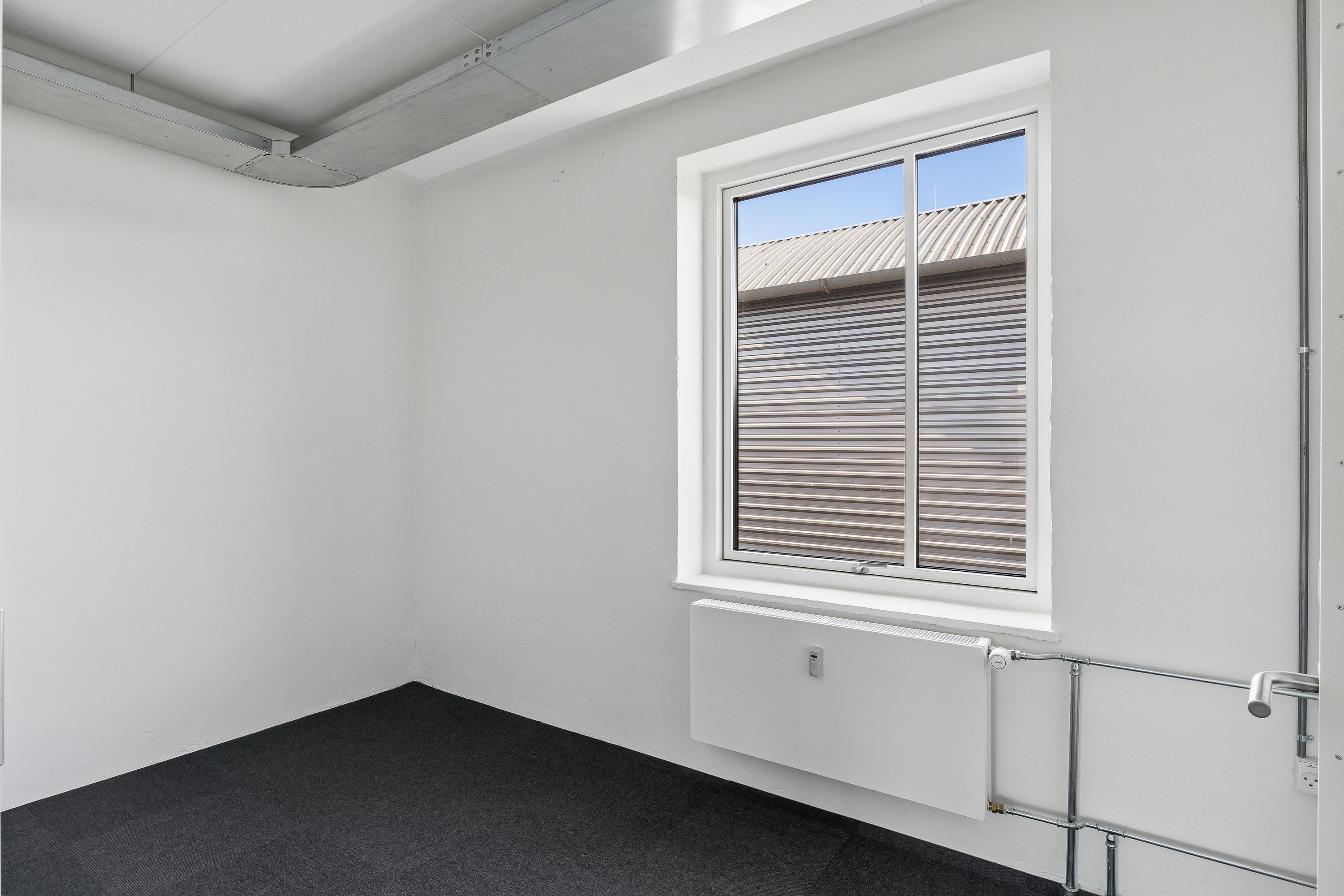 Lækkere kontorlokaler med hvidt loft og hvide vægge, mørke gulvbelægning og radiator. Vinduet med gardiner og udsigt til en bygning med stålgavl