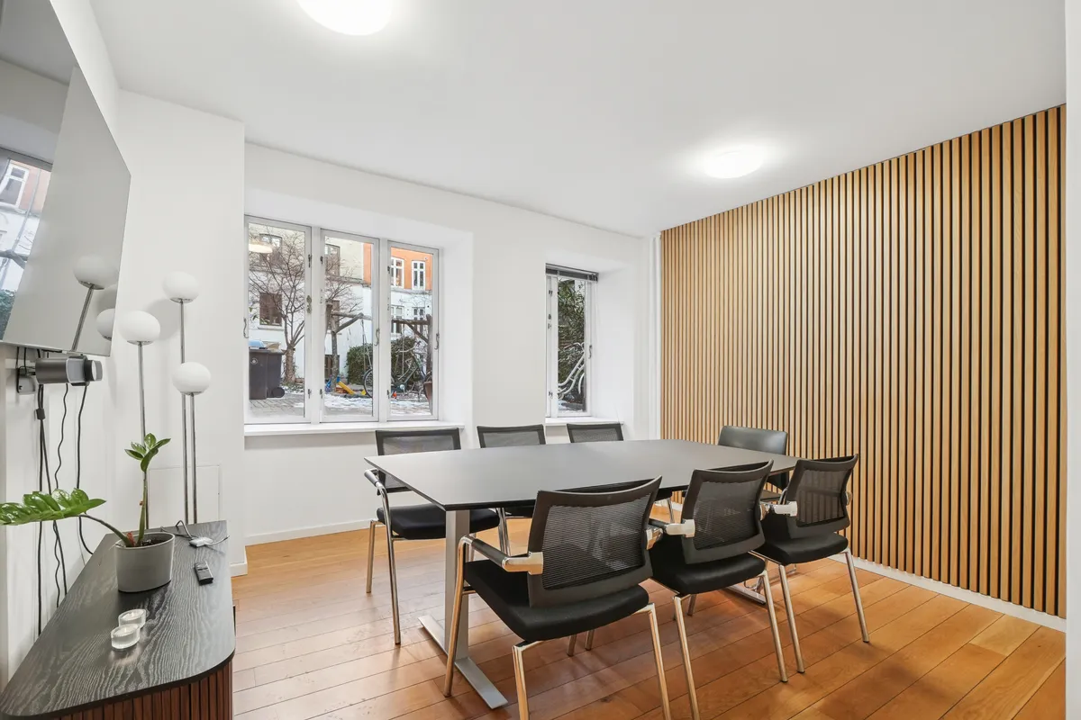 Minimalistisk mødelokale med højloft, bord og stole samt plante og lamper i baggrunden