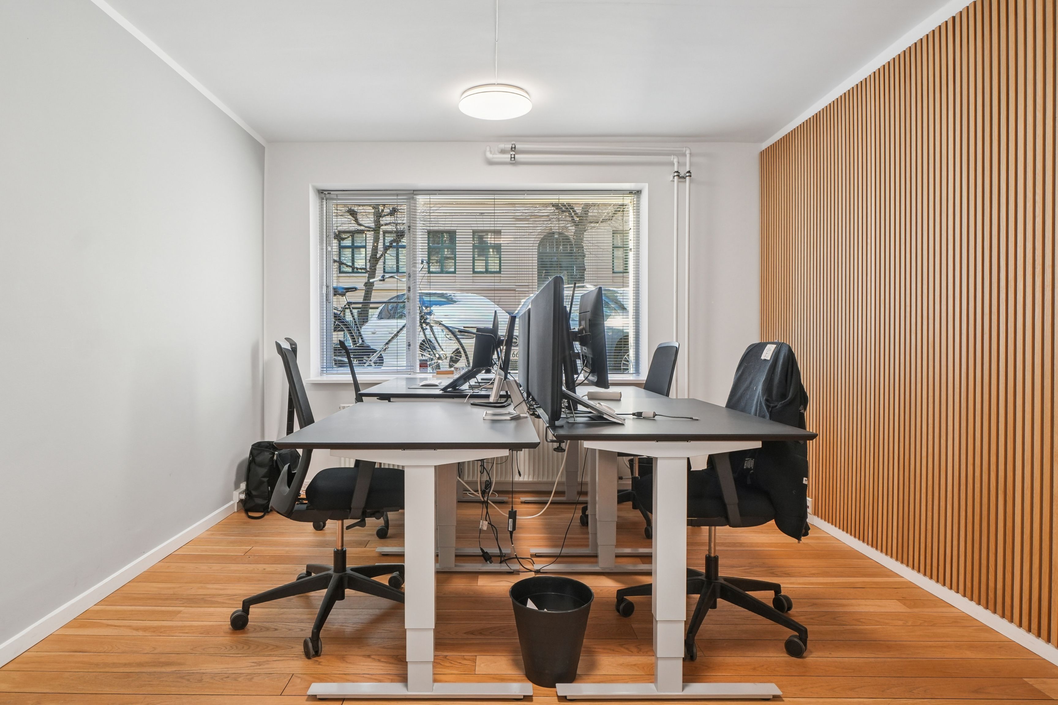 To kontorer med høje skriveborde og lyse gulv. To høje stole med ryglæn og hjul. To skærme og en sort papirkurv. Vindue med gardiner og udsigt til parkeringsplads