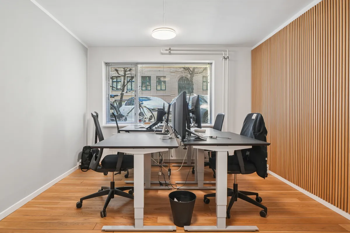 To kontorer med høje skriveborde og lyse gulv. To høje stole med ryglæn og hjul. To skærme og en sort papirkurv. Vindue med gardiner og udsigt til parkeringsplads