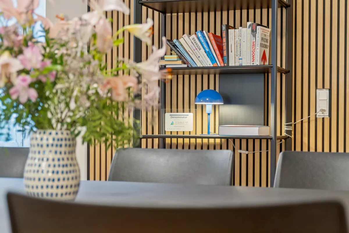 En moderne kontorrum med et bord og stole, en vase med blomster, en lille blå lampe og et hylde med bøger og en dekorative plakat