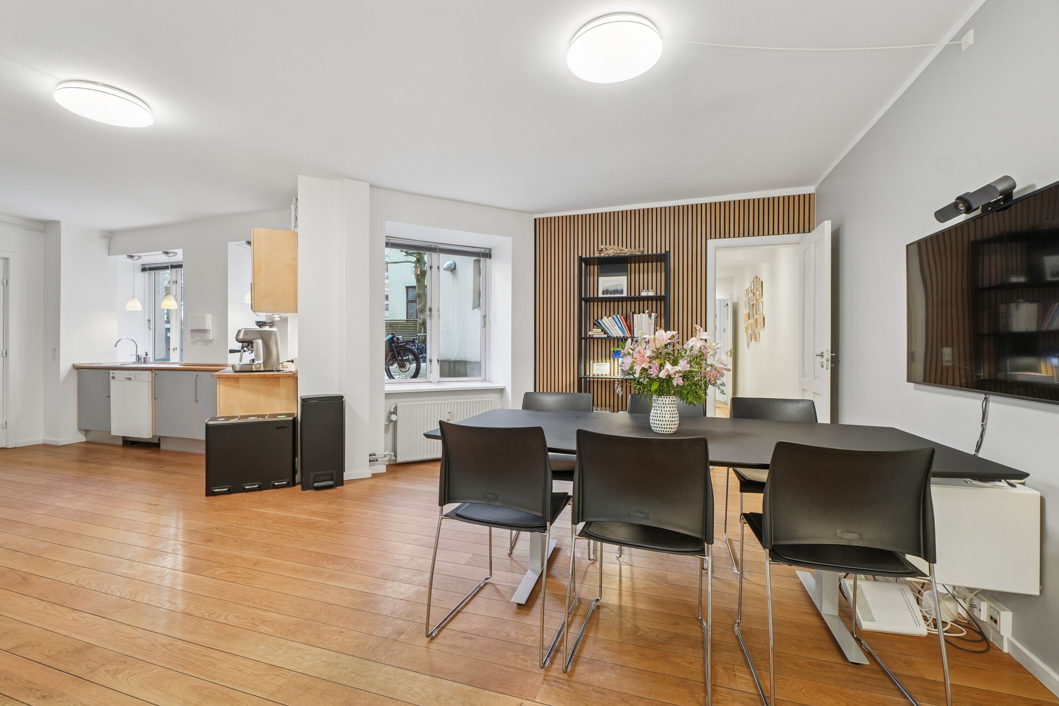 En moderne og lys kontor med bord og stole, lysekroner, blomster i vase, tv og en spisestue med køkken