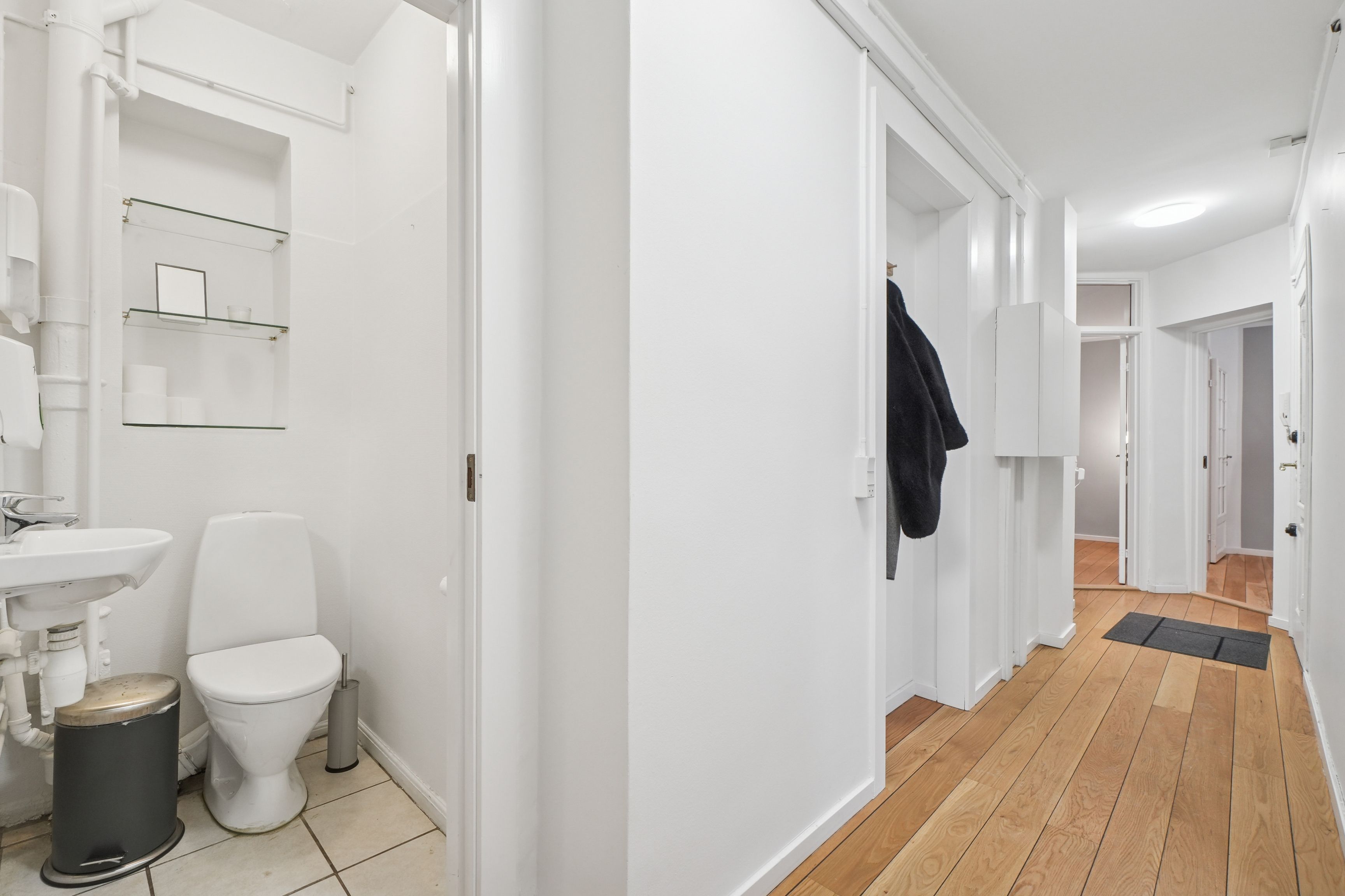 En hvid toilet med en bøjle hængende i toilettet og et toiletpapirhylde. Et hvidt badeværelse med et toilet, en hvid vask og et hvidt badeværelse.