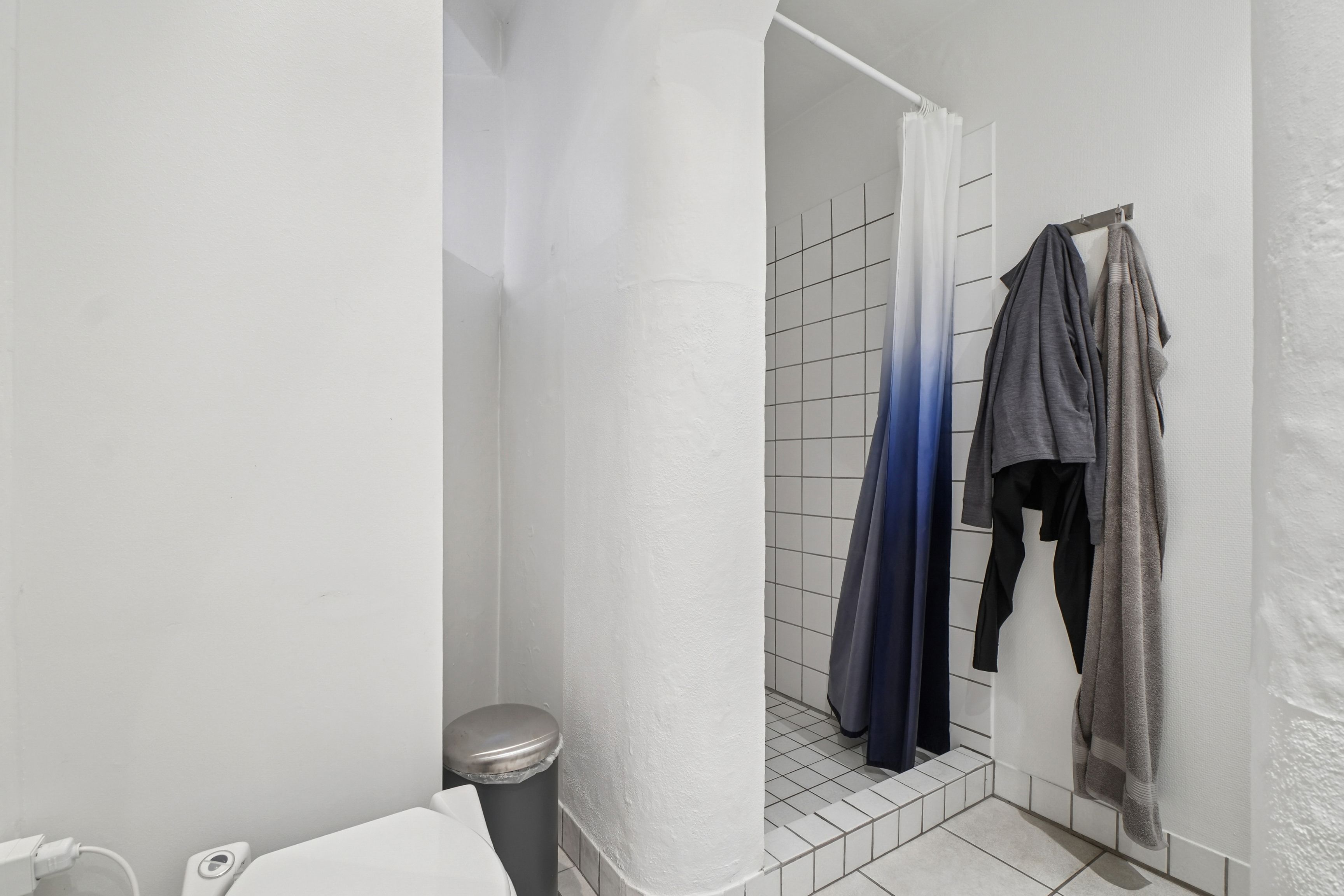 Et moderne badeværelse med hvid væg og hvidt toilet med toiletpapirholder samt en højt placeret hængekrog med tøj