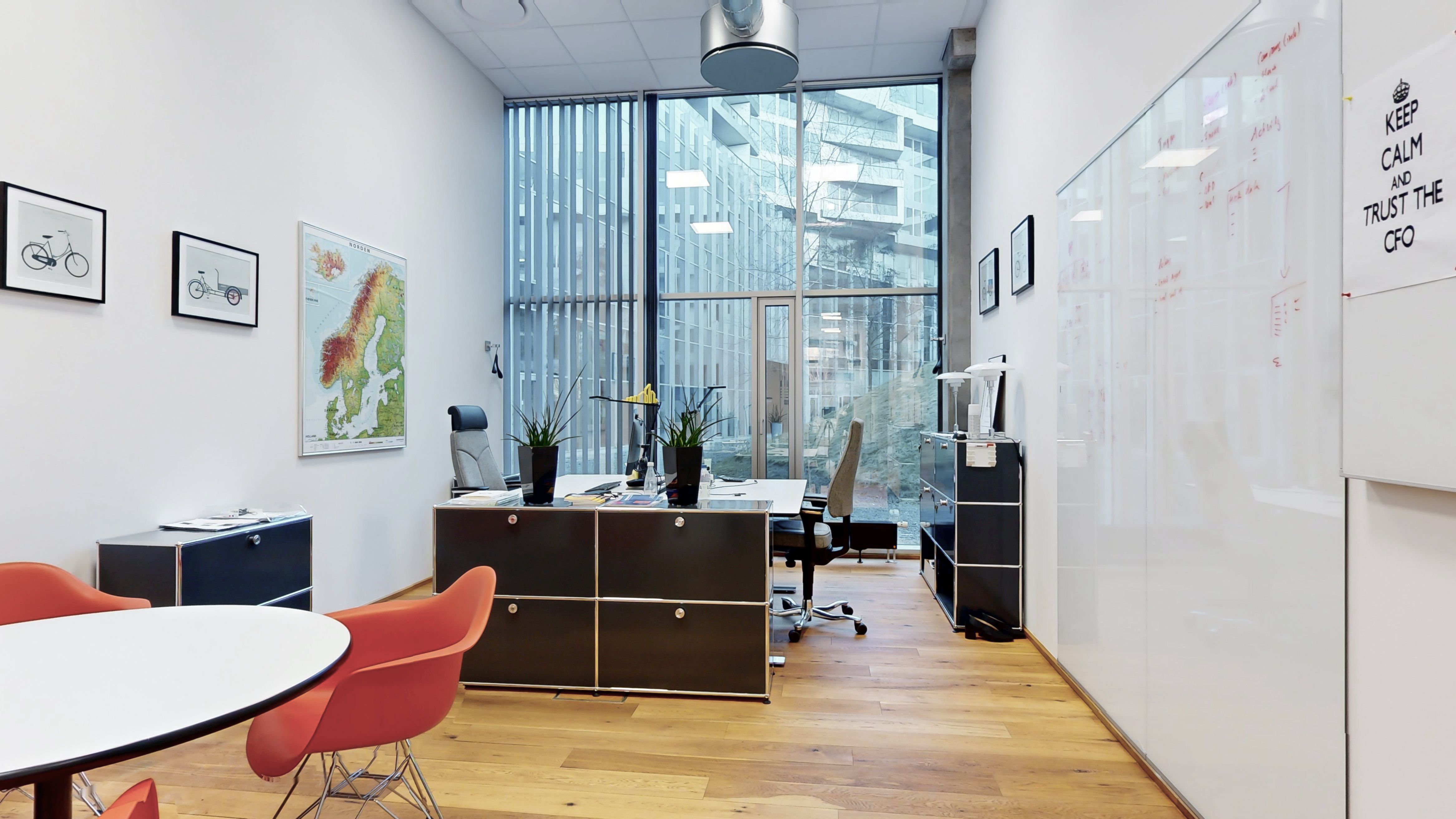 Billede af kontorlokaler med højt til loftet, store vinduer, moderne møbler og skrivebord. Væggene er hvide og har skrivebordsvægge. Der er kunstværker og planter på skrivebordene.