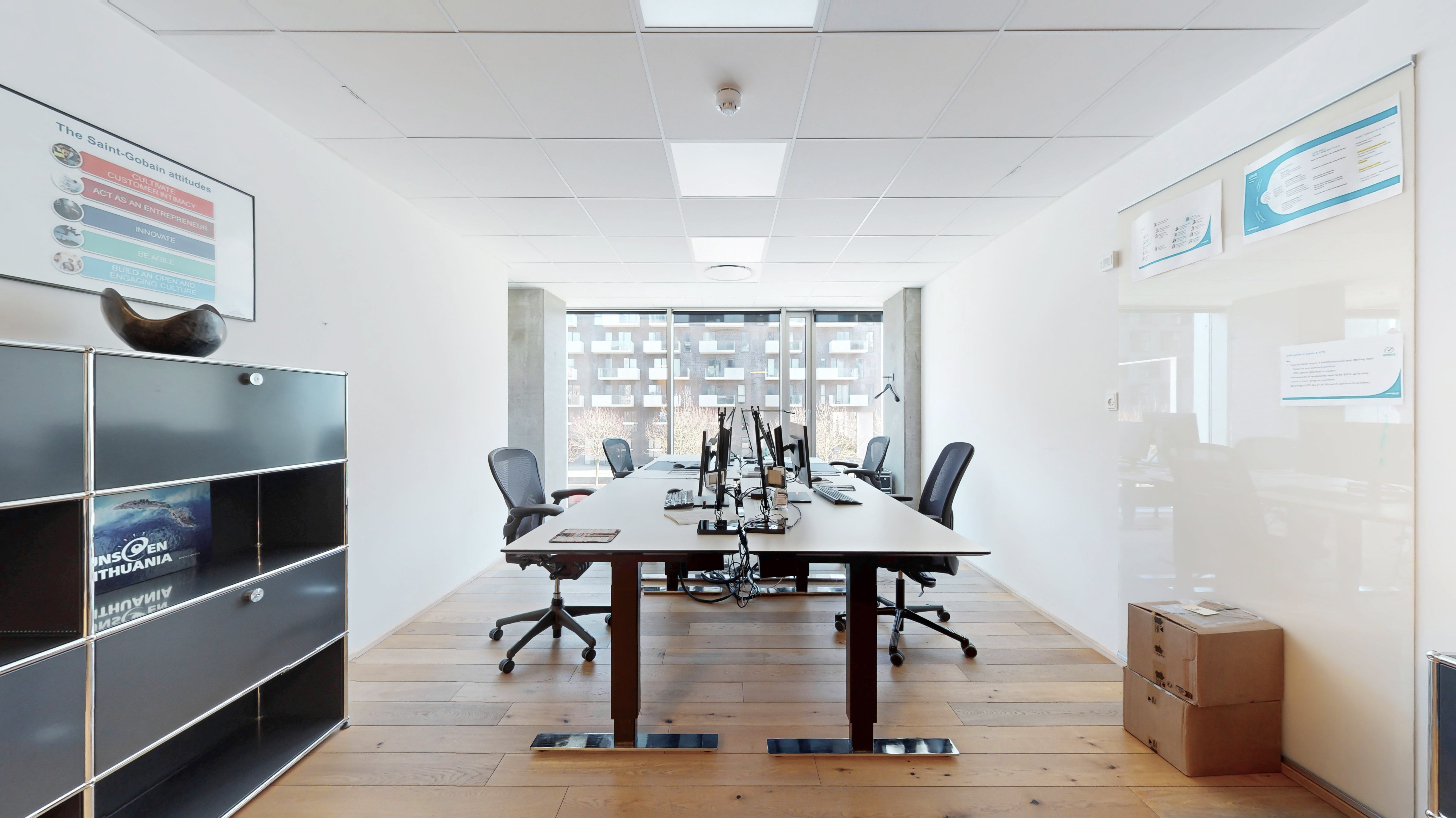 Et kontorlokal med lysegul væg og trægulv samt moderne møbler. Der er et skrivebord med computere og en stol. Der er også en høj kabinet og en væghængende plakat