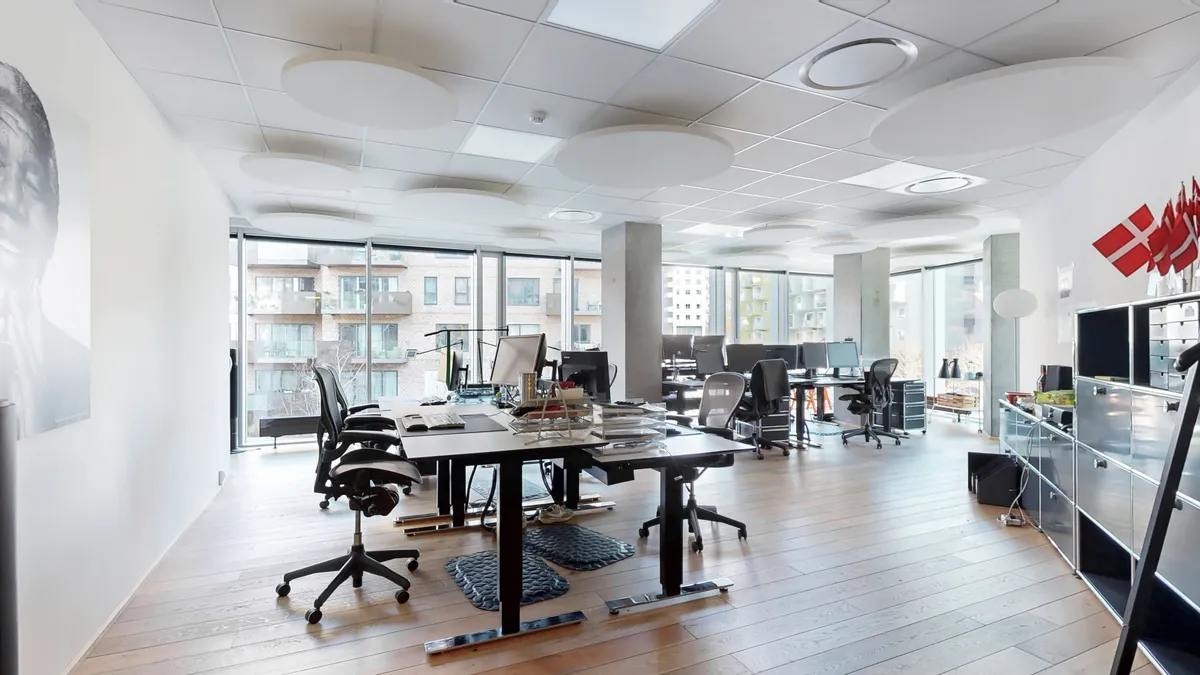 Moderne kontor med højt loft, lyse loftsbelægninger og stolpe med flag på væggen. Væggen er hvid og smukke trægulve. Møbler og computere er placeret i rummet