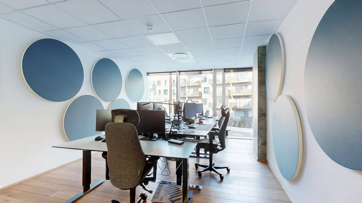 Moderne arbejdsplads med tre skriveborde, stol, computere, og runde gulve. Store vinduer med udsigt til et byggeprojekt.