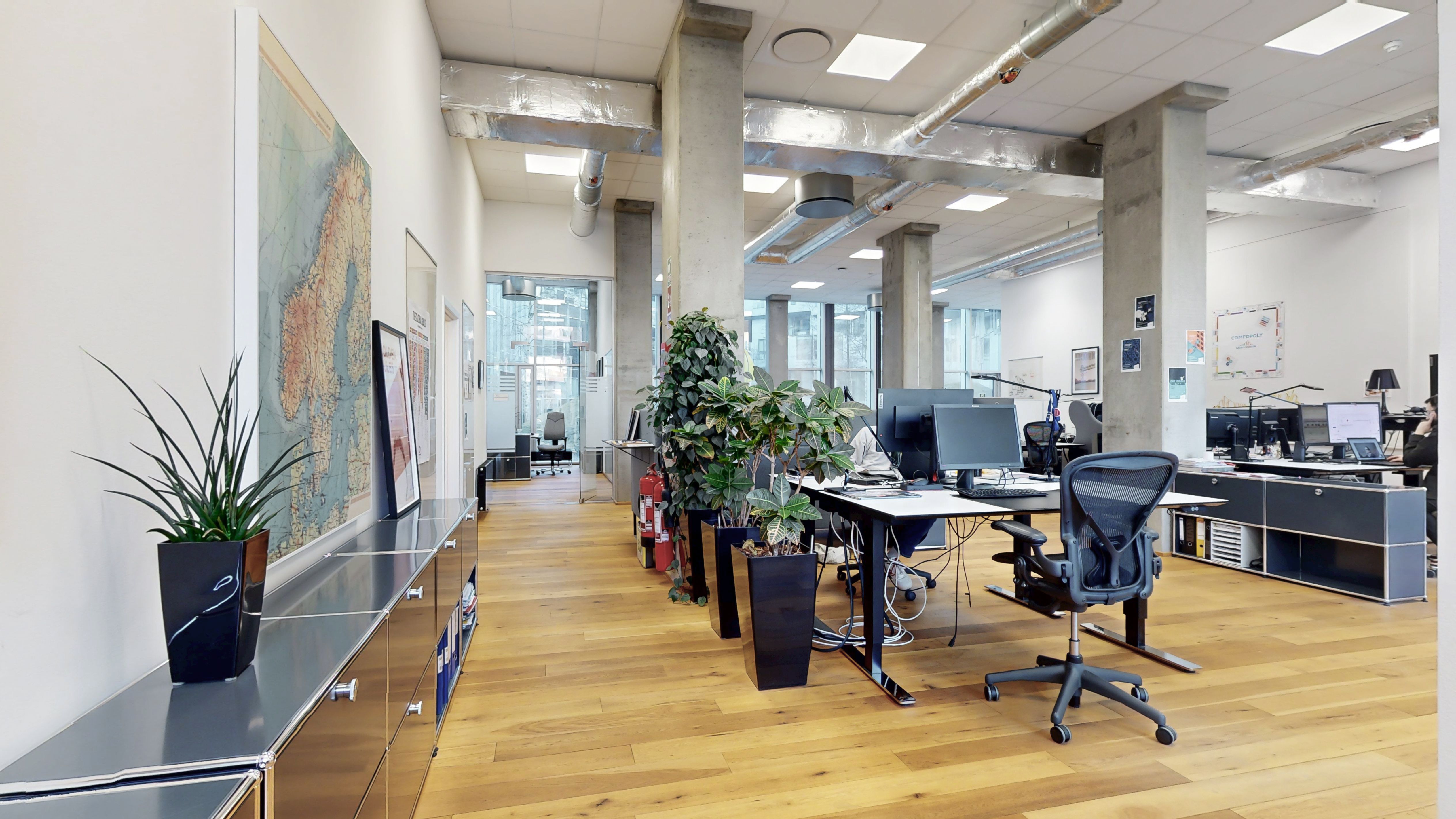 Et moderne kontor med højt til loftet og lyse loftsbrædder. Bordplader med computere og kontorstolen og planterne er placeret i midten af lokalet.