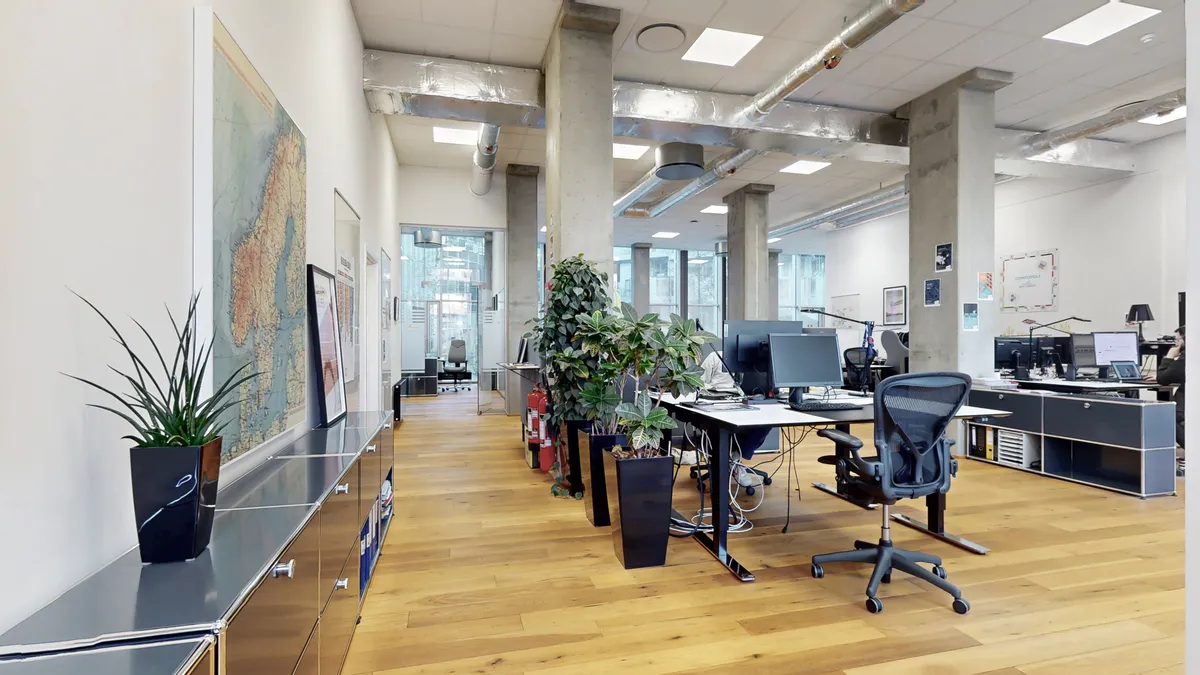Et moderne kontor med højt til loftet og lyse loftsbrædder. Bordplader med computere og kontorstolen og planterne er placeret i midten af lokalet.