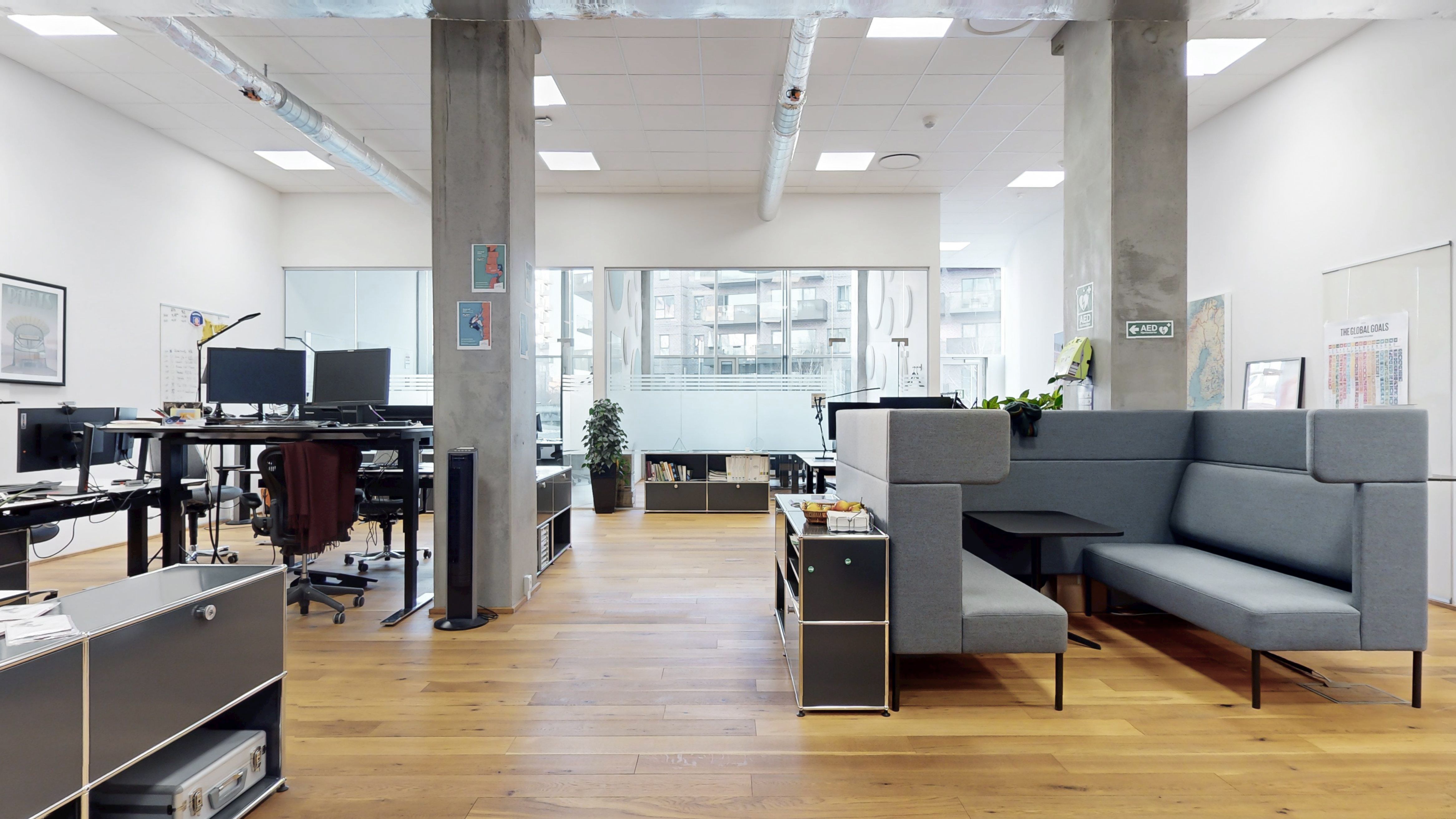 Kontorrum med skriveborde, stole og plante i hjørnet. Bord med skærme og skriveborde. To lænestol i gråt læder. En lænestol med sidebord og frugtkurv.