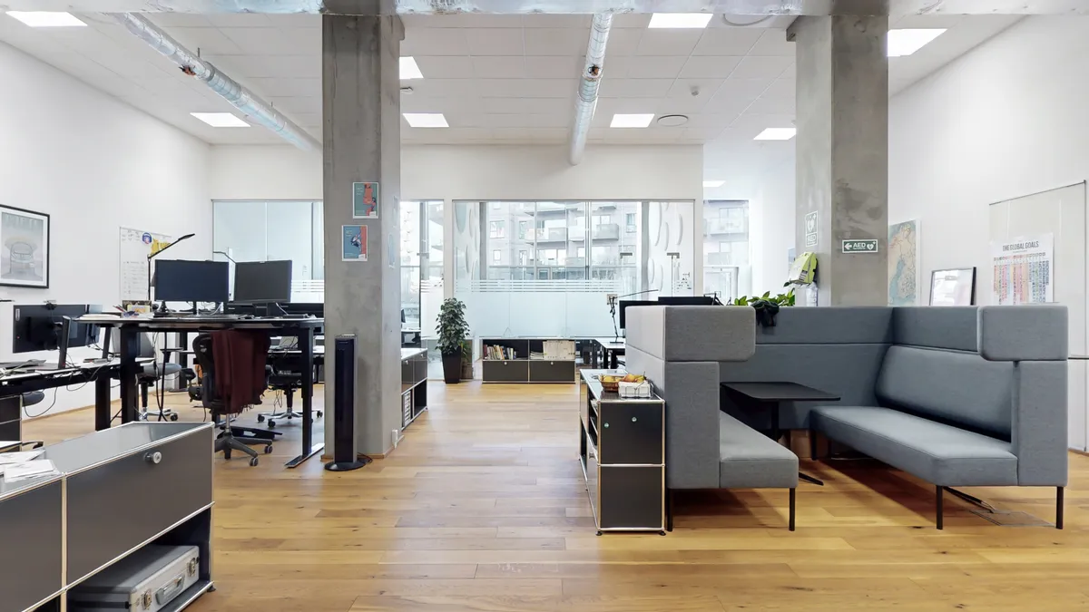 Kontorrum med skriveborde, stole og plante i hjørnet. Bord med skærme og skriveborde. To lænestol i gråt læder. En lænestol med sidebord og frugtkurv.