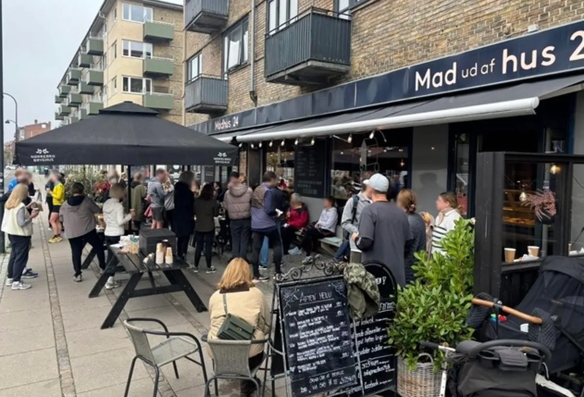 En stor mængde mennesker venter i kø foran Mad ud af hus på Søborg Hovedgade. Kunder nyder deres morgenmad udenfor restauranten og nyder selskab. Der er mange folk på gågaden, og der er en stor mængde grønne planter på siden.