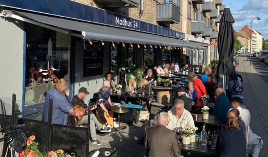 Mange gæster sidder og spiser på restauranten Madhus 24 i Dyssegård. Musikere spiller foran dem, og gæsterne ser ud til at nyde mad og musik.