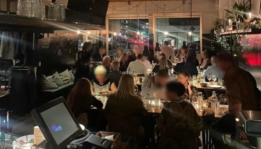 Mange mennesker sidder og spiser på restauranten med borde, der er fulde af vin og øl. Der er også en kasseapparat og en monitor på en af bordene.