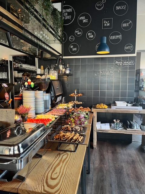 Buffet med frisk frugt og kødretter på en træbord, en flaske og en plante i baggrunden og et sort vægmalerier med skrift og lysereflekser