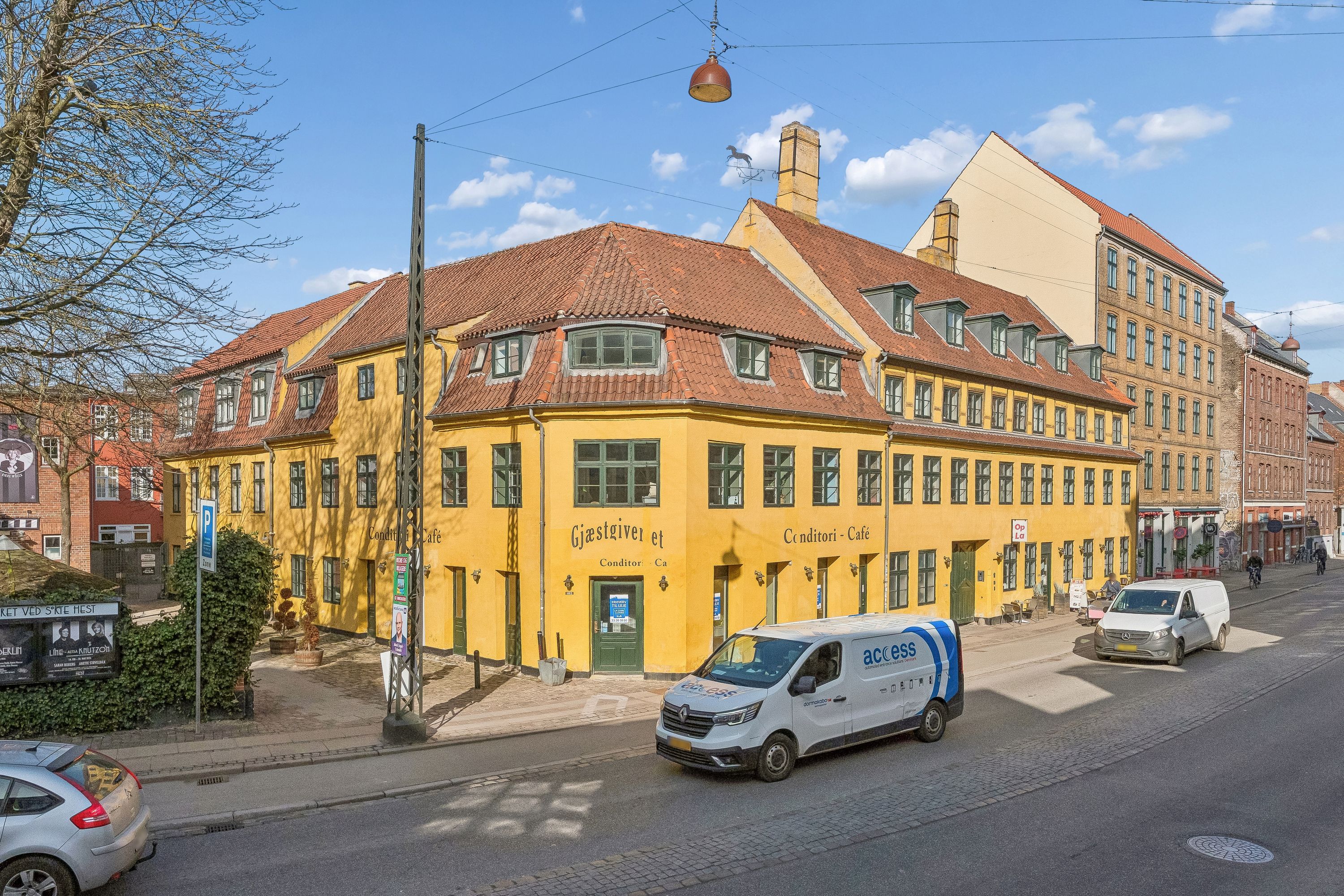 To kørende køretøjer, en bil og en van, kører ad en tosporet vej. Der er en cafe med navnet Gjæstgiveri et Conditori Cafe. Der er en stående cykelkører og et schild med en mand og tekst på en bygning