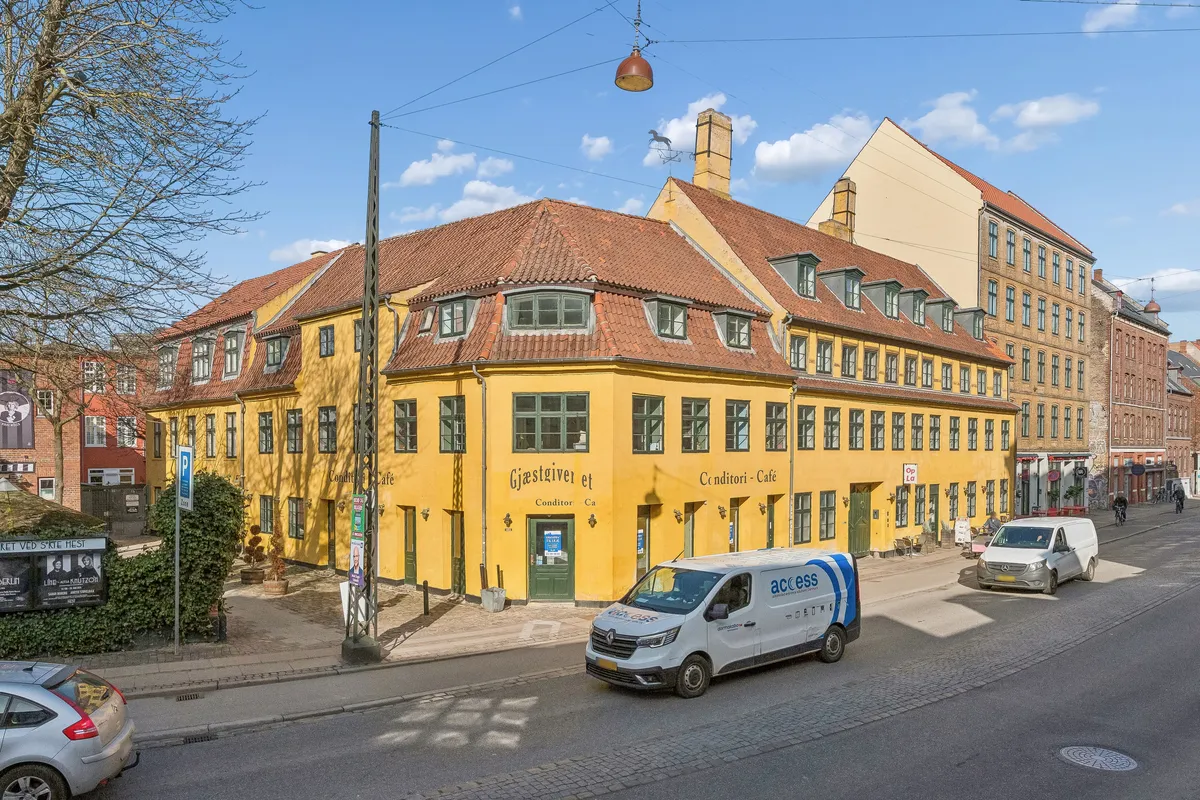 To kørende køretøjer, en bil og en van, kører ad en tosporet vej. Der er en cafe med navnet Gjæstgiveri et Conditori Cafe. Der er en stående cykelkører og et schild med en mand og tekst på en bygning