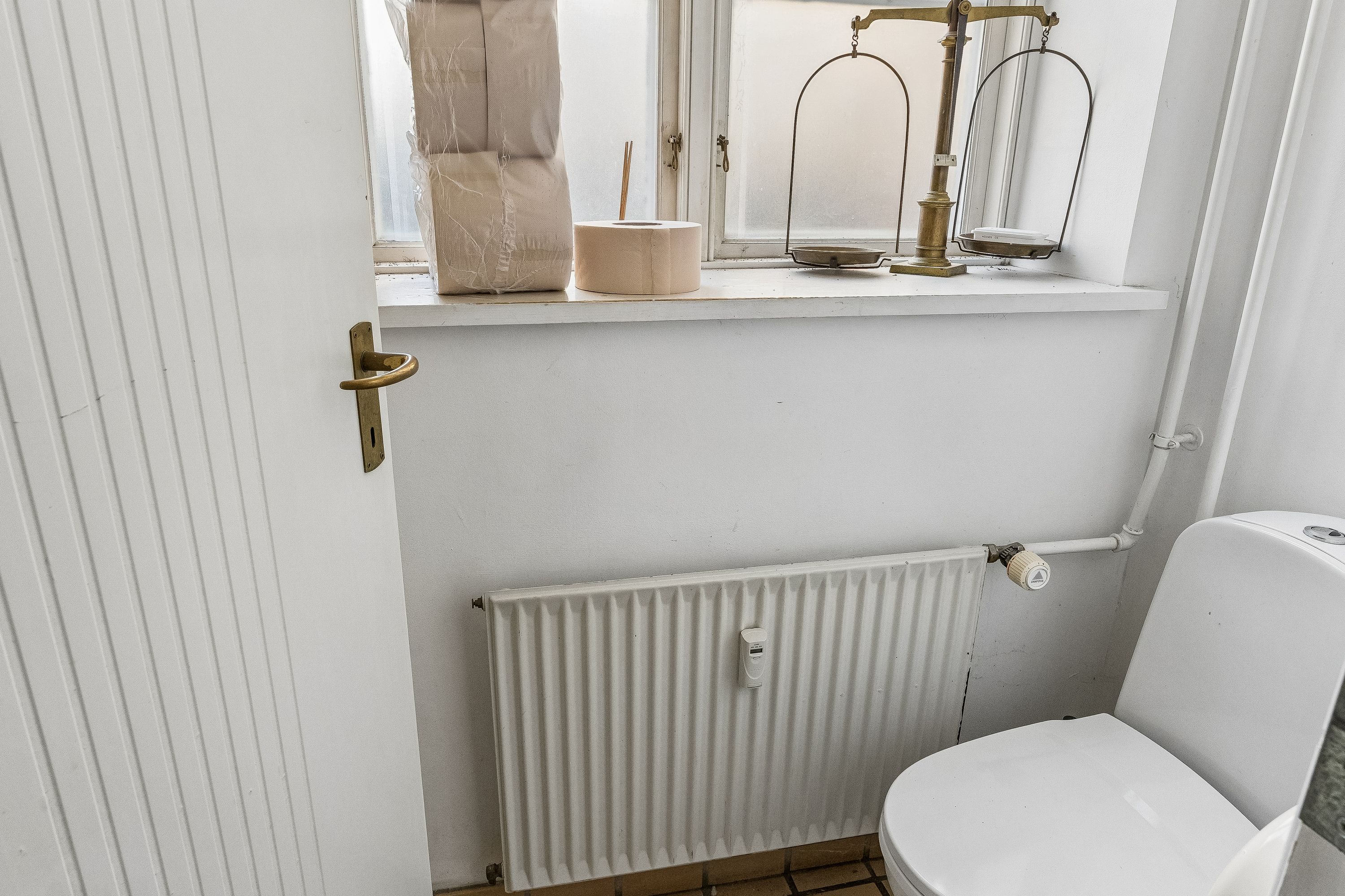 En lille toilet med radiator, der står i et rum med hvide vægge og et vindue med to papkasser og toiletpapir