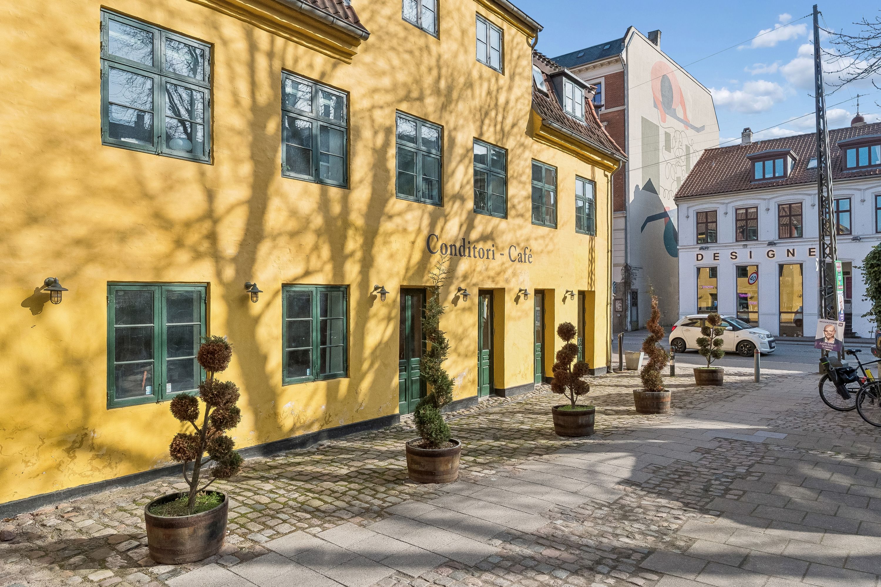En gul bygning med butikker og cafeer med toppede potteplanter på fortovet og en bil parkeret i baggrunden