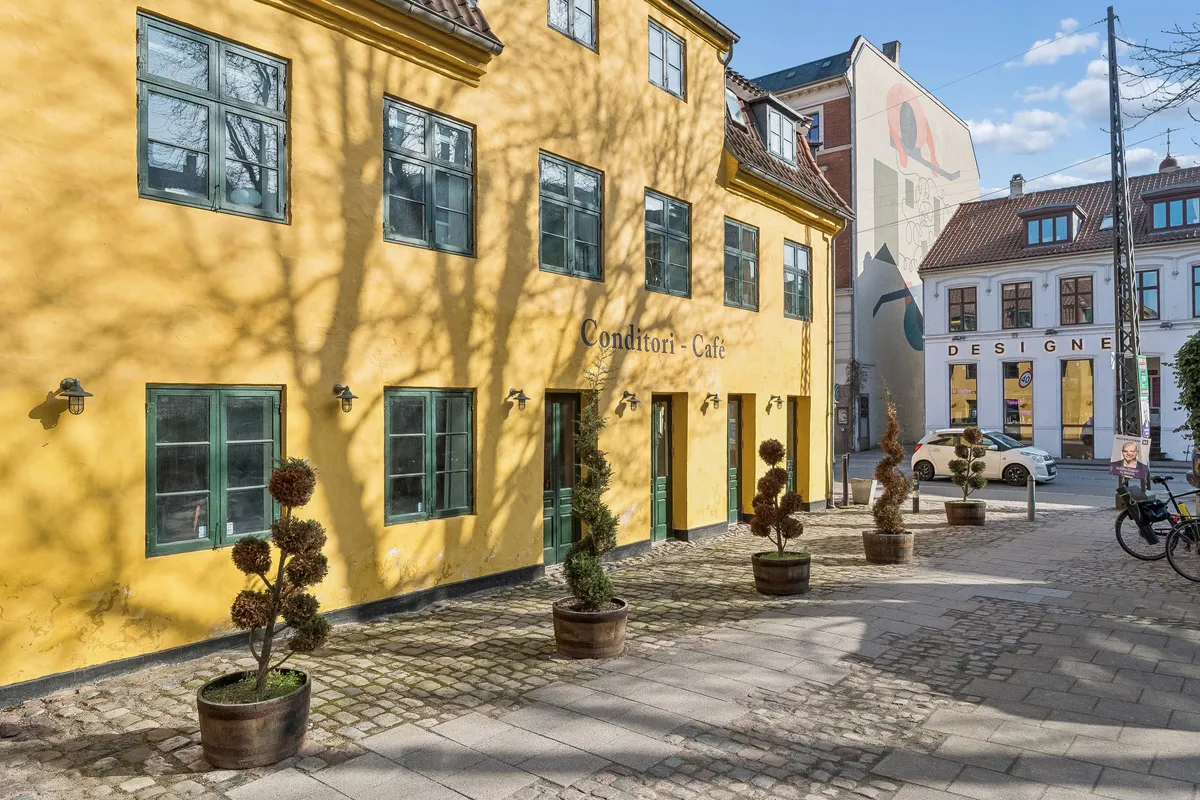 En gul bygning med butikker og cafeer med toppede potteplanter på fortovet og en bil parkeret i baggrunden