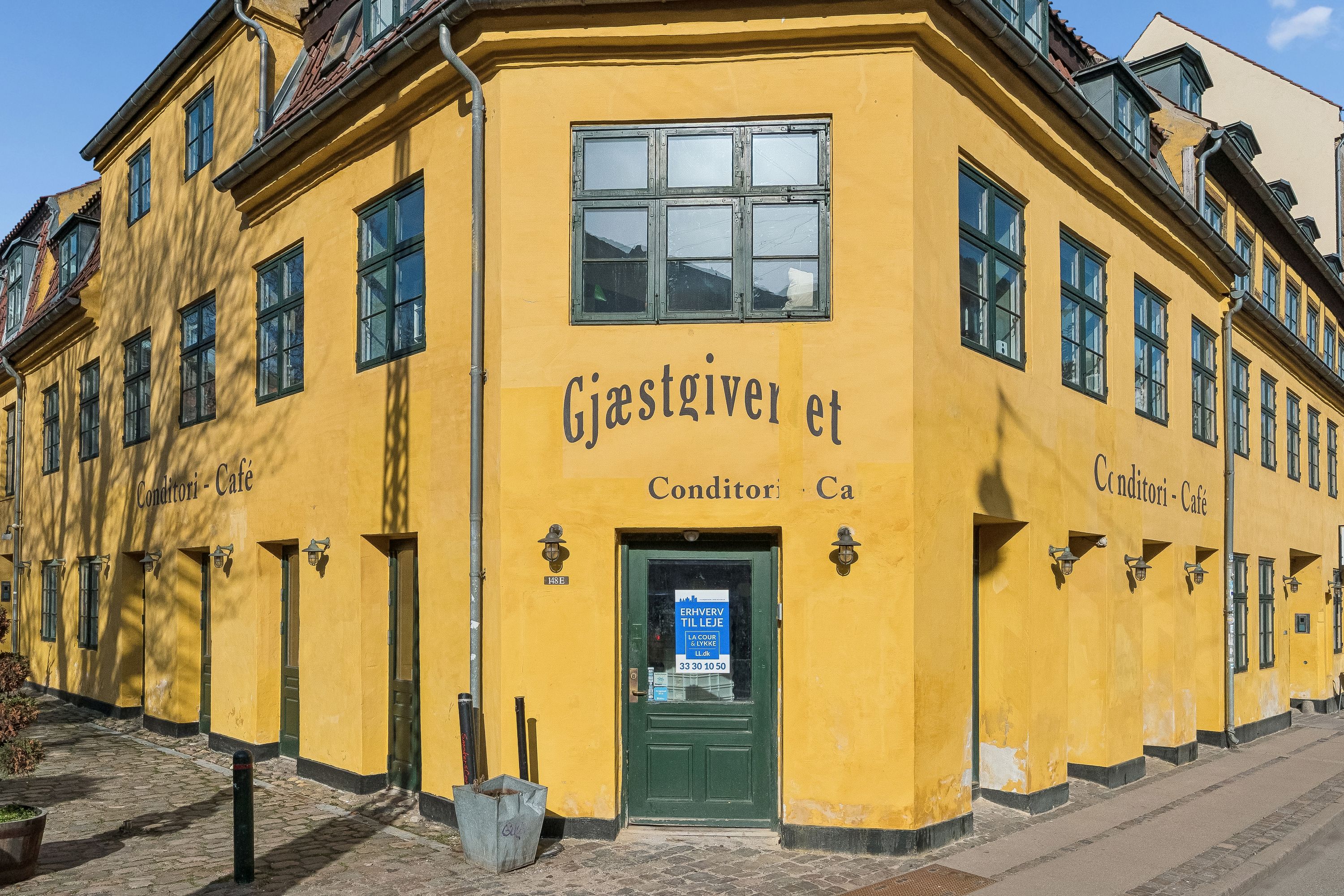 Gul bygning med rødt tag, beliggende på Vesterbrogade 148E, København V. Målskilt med teksten Gjæstgiveren et Conditori Ca. Gule vægge med grønne ruder og lysekroner. En grøn dør med et vinduesskilt og et skovl