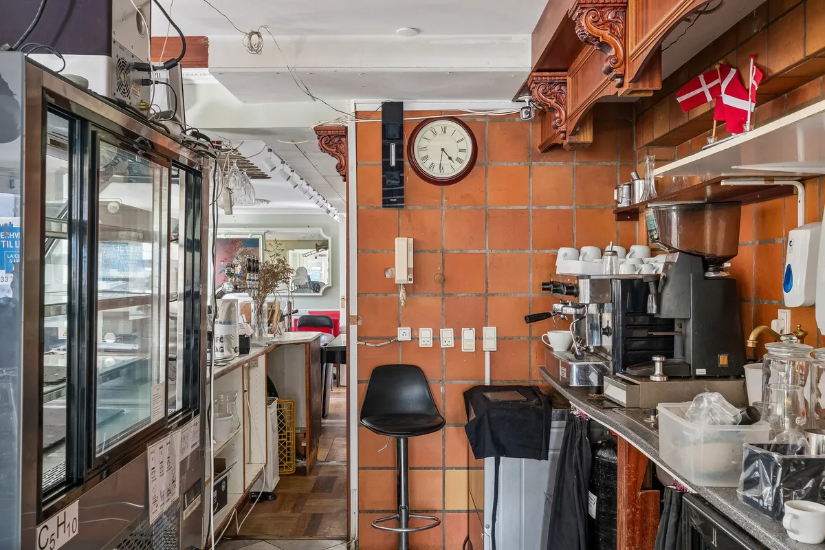 Køkken med kaffemaskine og urtrækning, ordnede hylder, gul urtavle og små flag i baggrunden, smukt oplyst af lysekroner og lyse gulve