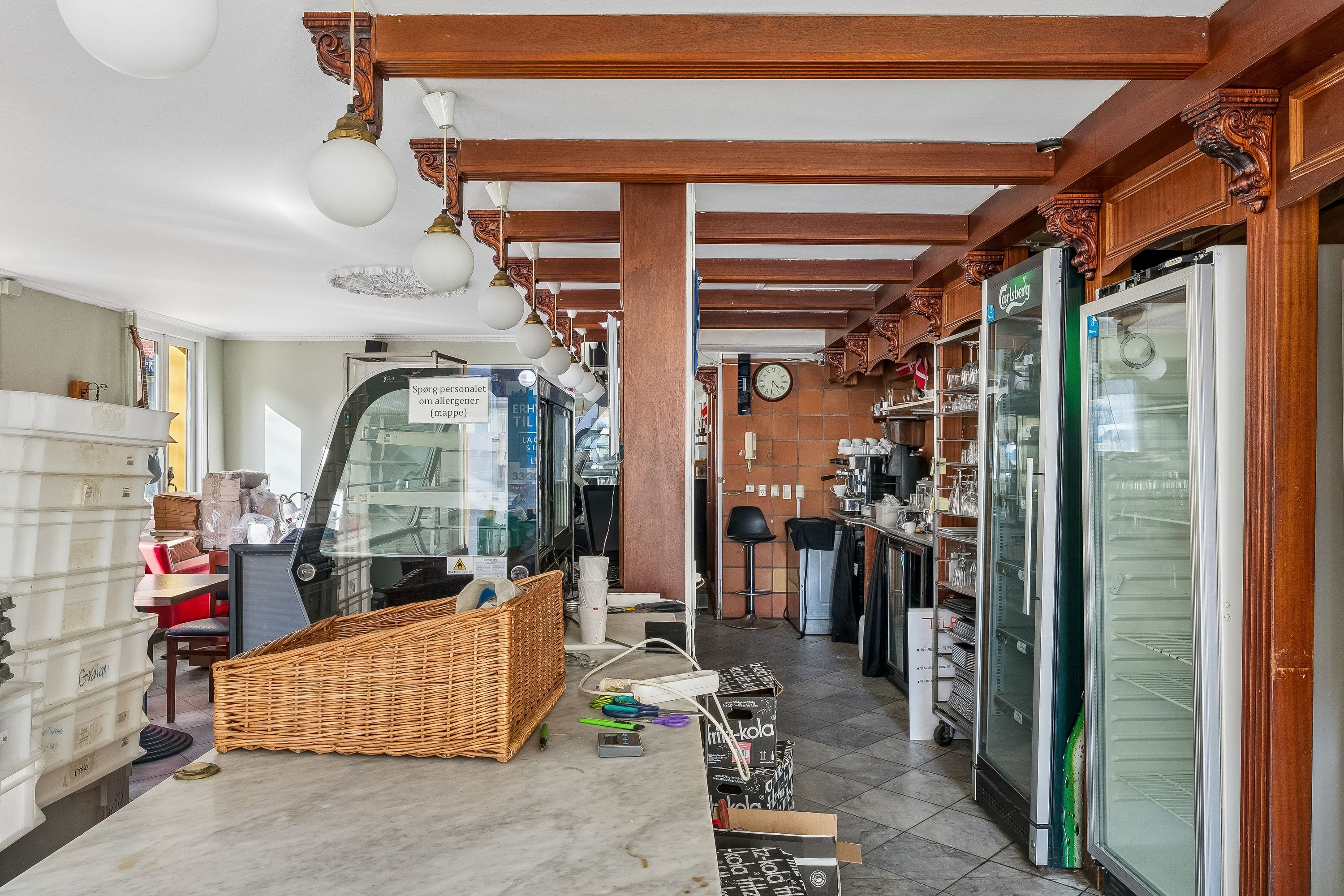 Indvends i caféen ses et køkken med en kobberplade, ølkøleskabe, en bordplade med papkasser og en kurv, lysekroner og en klokkeskærm