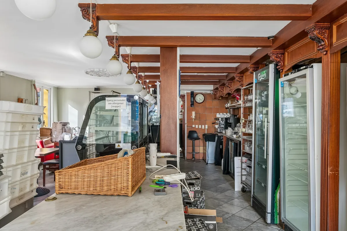 Indvends i caféen ses et køkken med en kobberplade, ølkøleskabe, en bordplade med papkasser og en kurv, lysekroner og en klokkeskærm