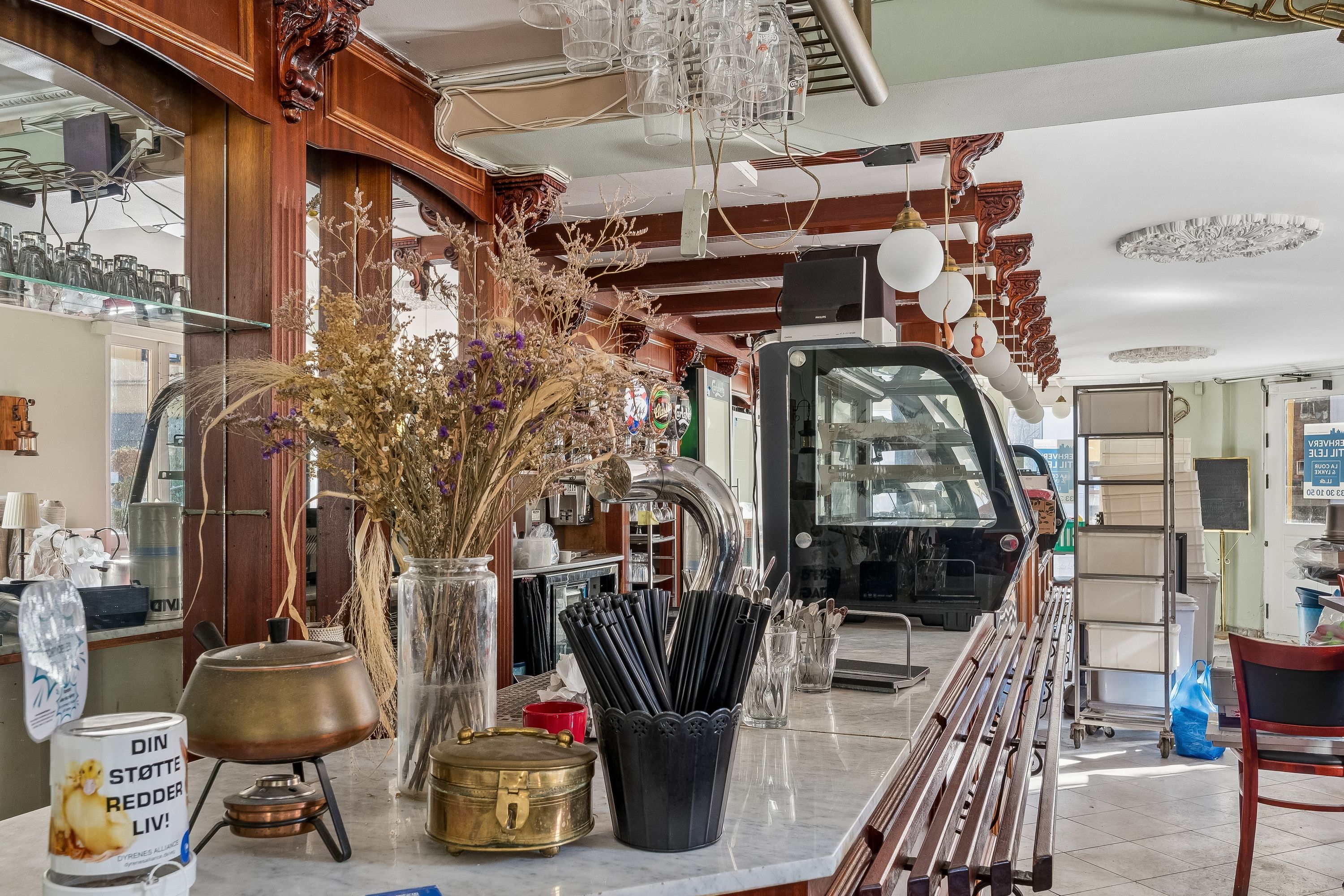 Café med marmorbord og stole, krydrede grøntsager og blomster i vase, glas og køkkenudstyr i baggrunden, samt en åben dør og vinduer med udsigt til gaden