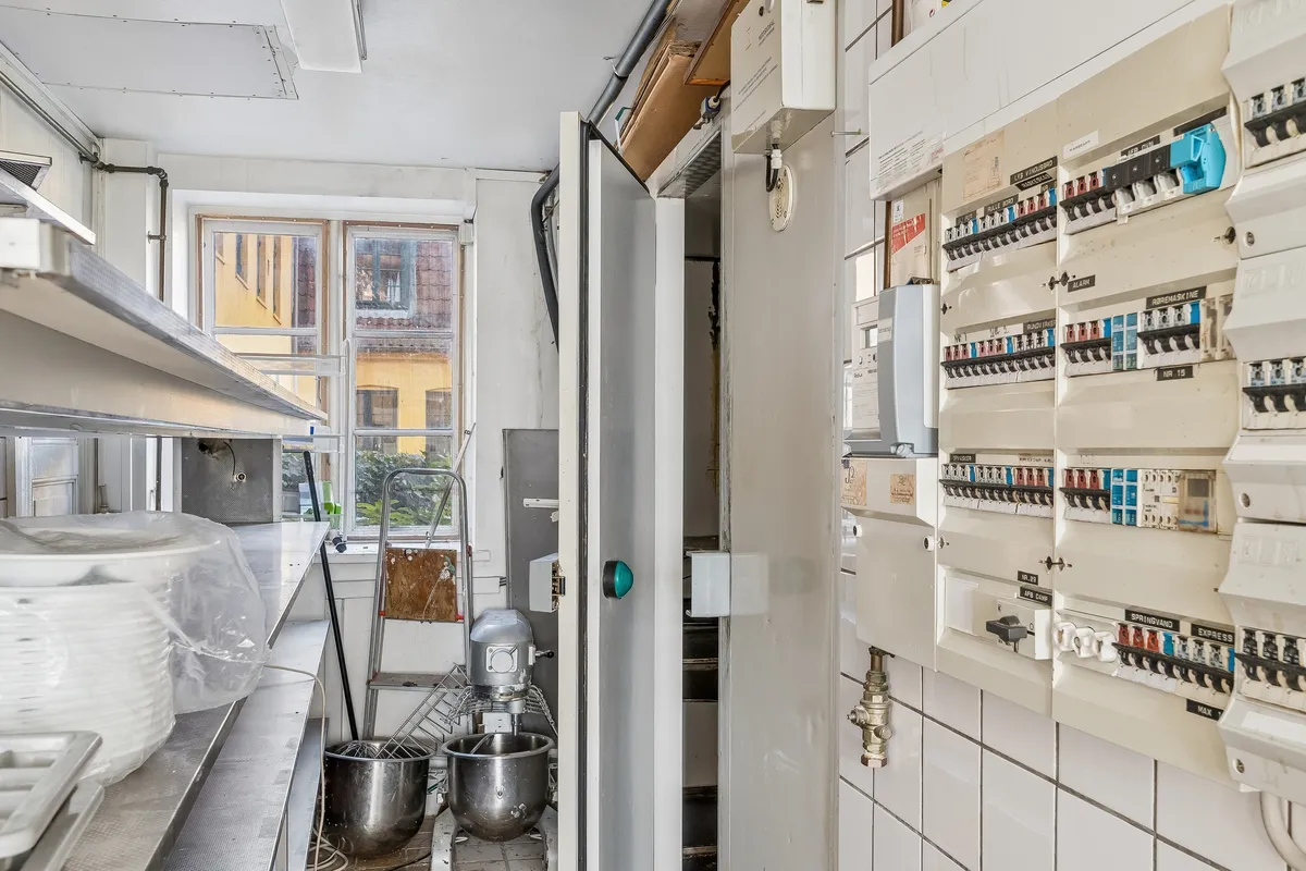 Køkken med komfur, blander og højre elektriske slukkontakter og tæller. To store stålbøjler med små højttalere og bøjle med plastikposer på loftet.