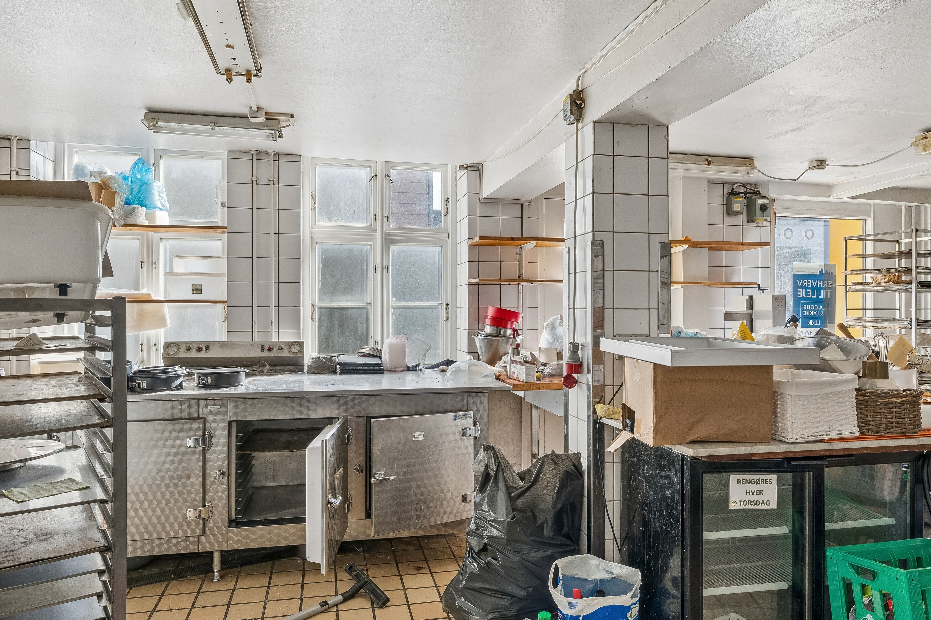 Køkken med ovn, komfur, skraldespand og opbevaringsrum. Køkkenbord med kasser og søndagsavis. Trækasser og metalhylder. Tidligere bageri.