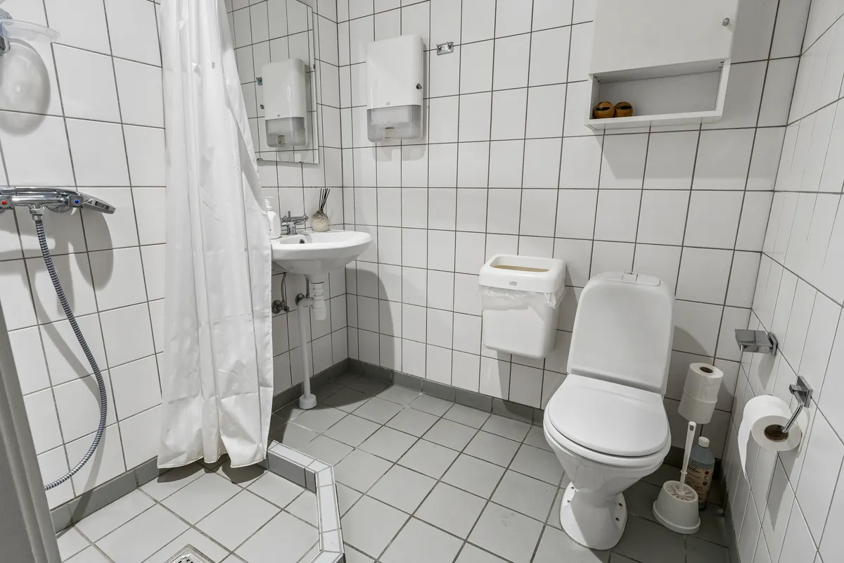 Bad med hvidt keramik toilet, hvidt vask og hvidt bruserhylde. Hvidt håndklædehylde og to toiletpapirruller. Spejl og håndklædetørrer i badeværelset.