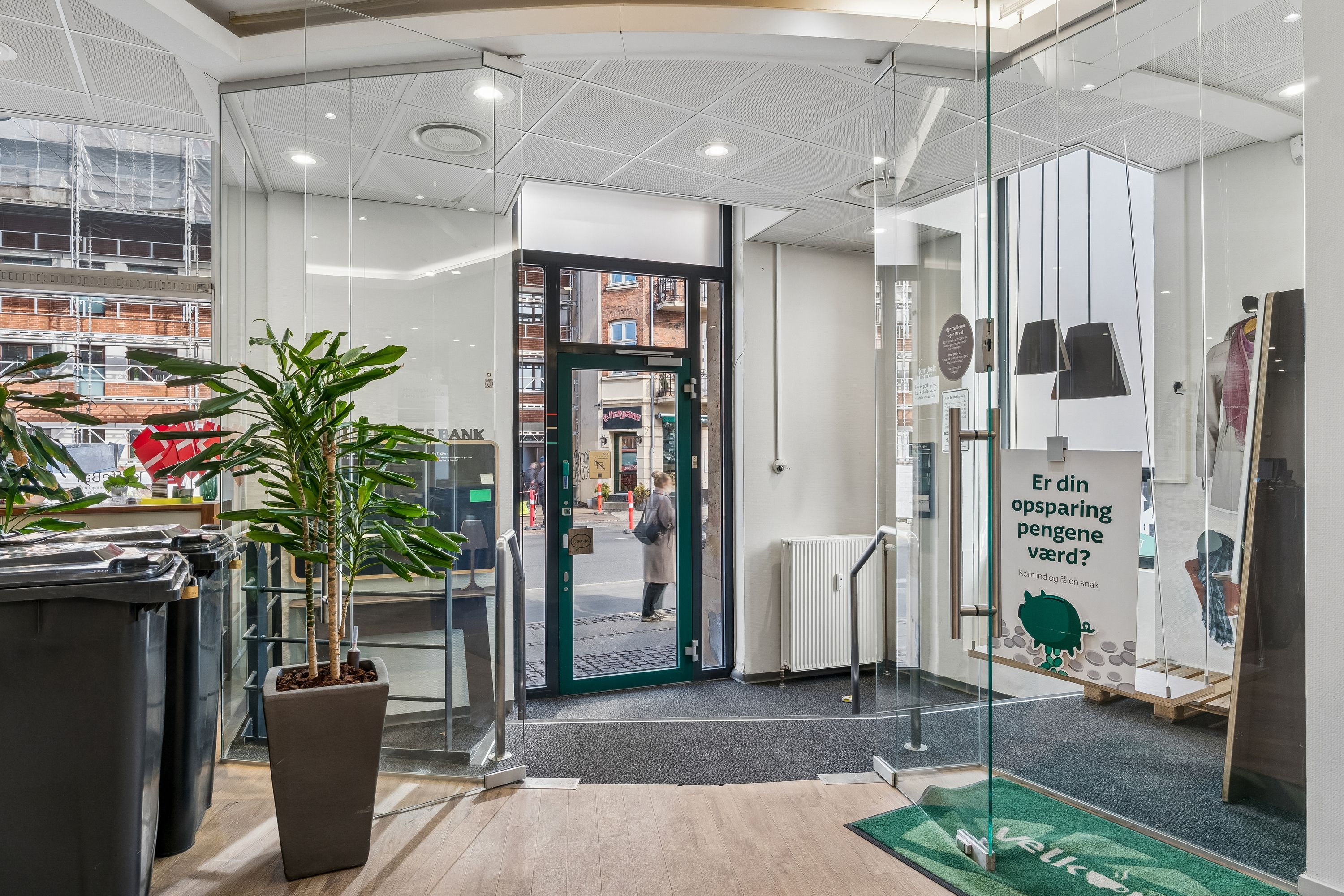 Kontor med glasvægge og grønne planter. Et smilt skilt med spørgsmålet: Er din opsparing pengeværd? En kvinde går ind gennem en grøn dør.